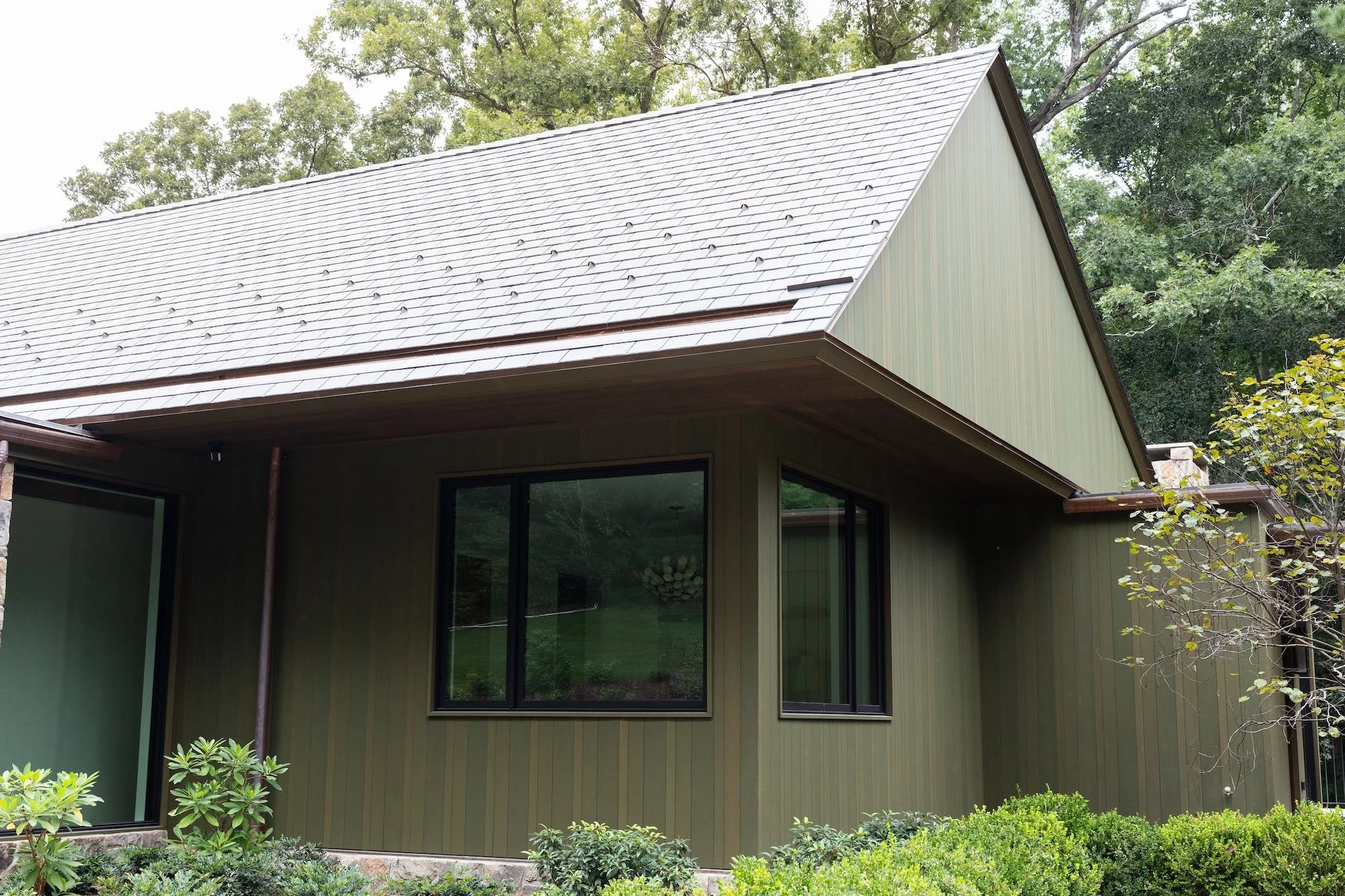 Modern house with green vertical siding, large black-framed windows, gabled roof with gray shingles, surrounded by foliage and trees.
