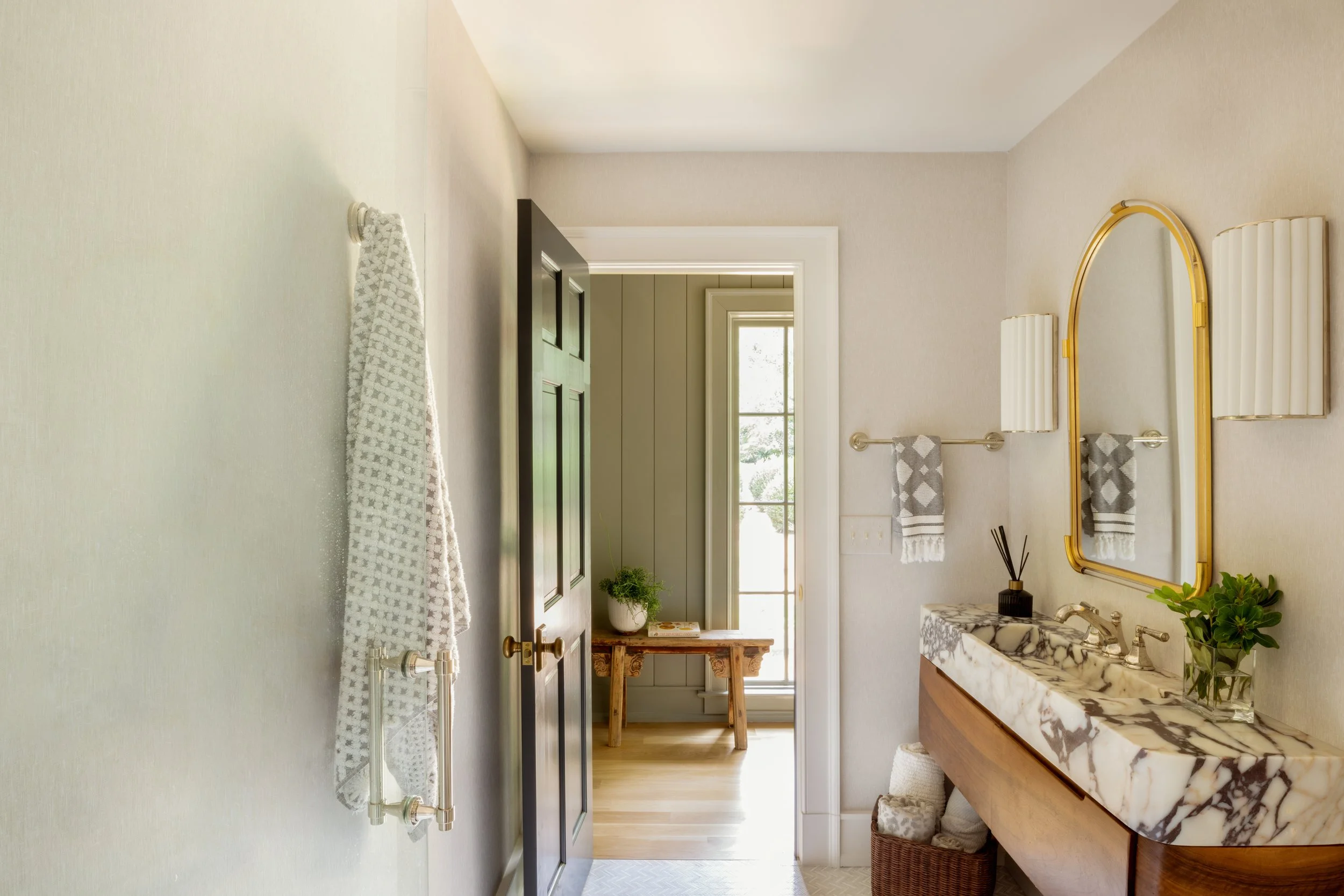 Interior view of a bathroom with a marble countertop sink, a large golden mirror, and decorative towels, with a door open revealing a sunlit room with a window and a small wooden table with plant and books.