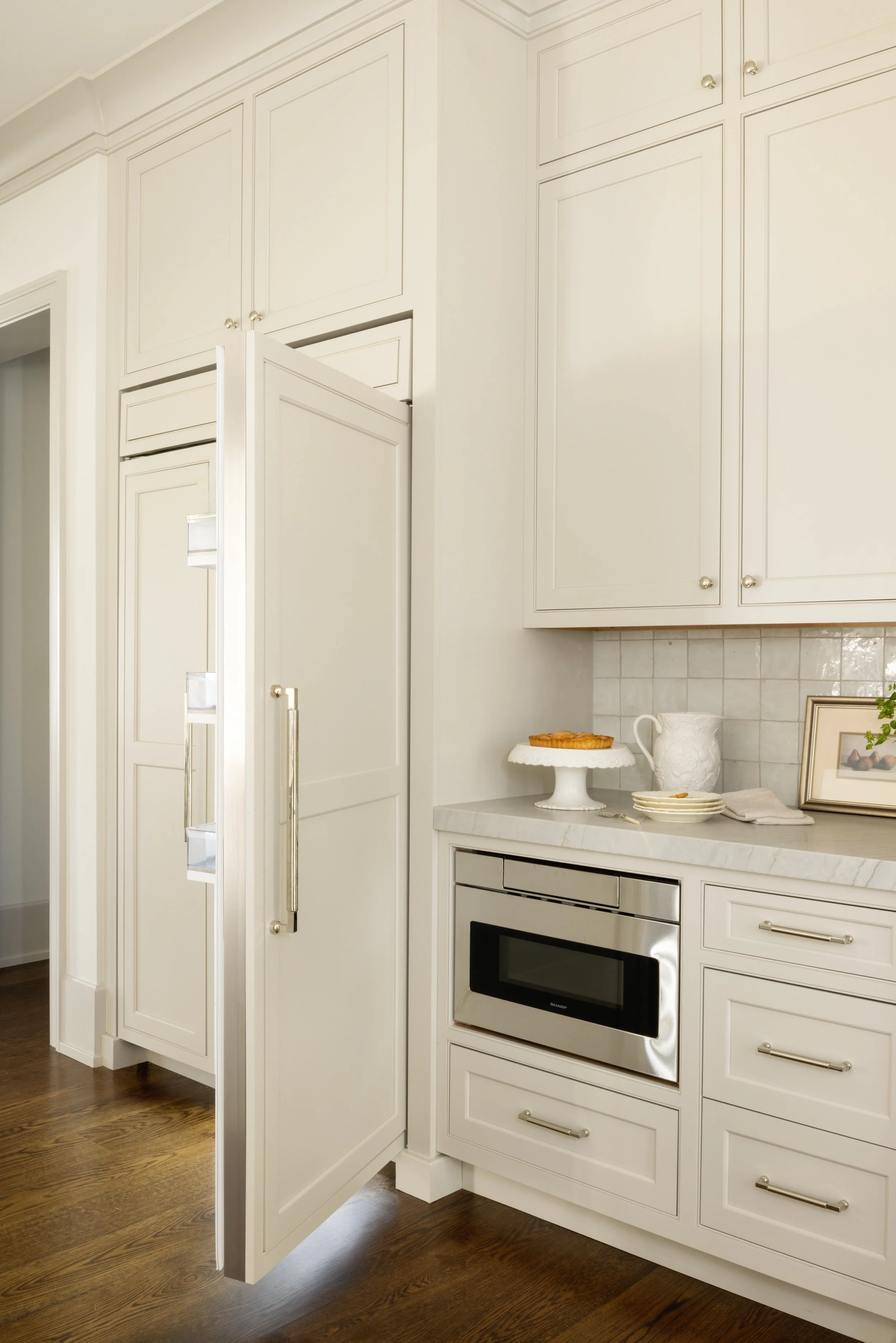 A white kitchen corner with cabinetry, a built-in microwave, a marble countertop, and decorative items like a pie on a stand, a white pitcher, and a framed photo.