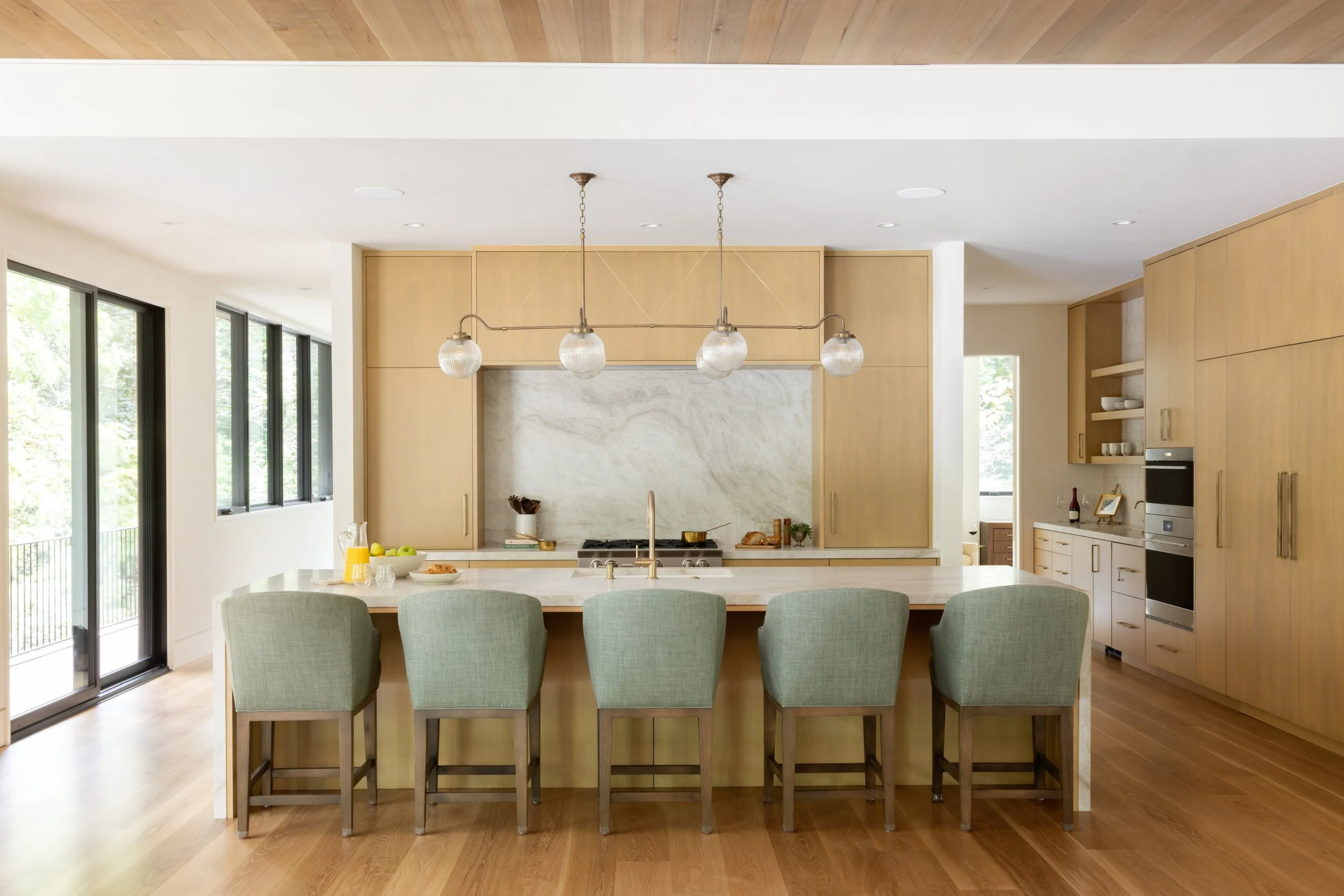 Modern kitchen with light wood cabinets, a white marble island with green upholstered chairs, and large windows letting in natural light.
