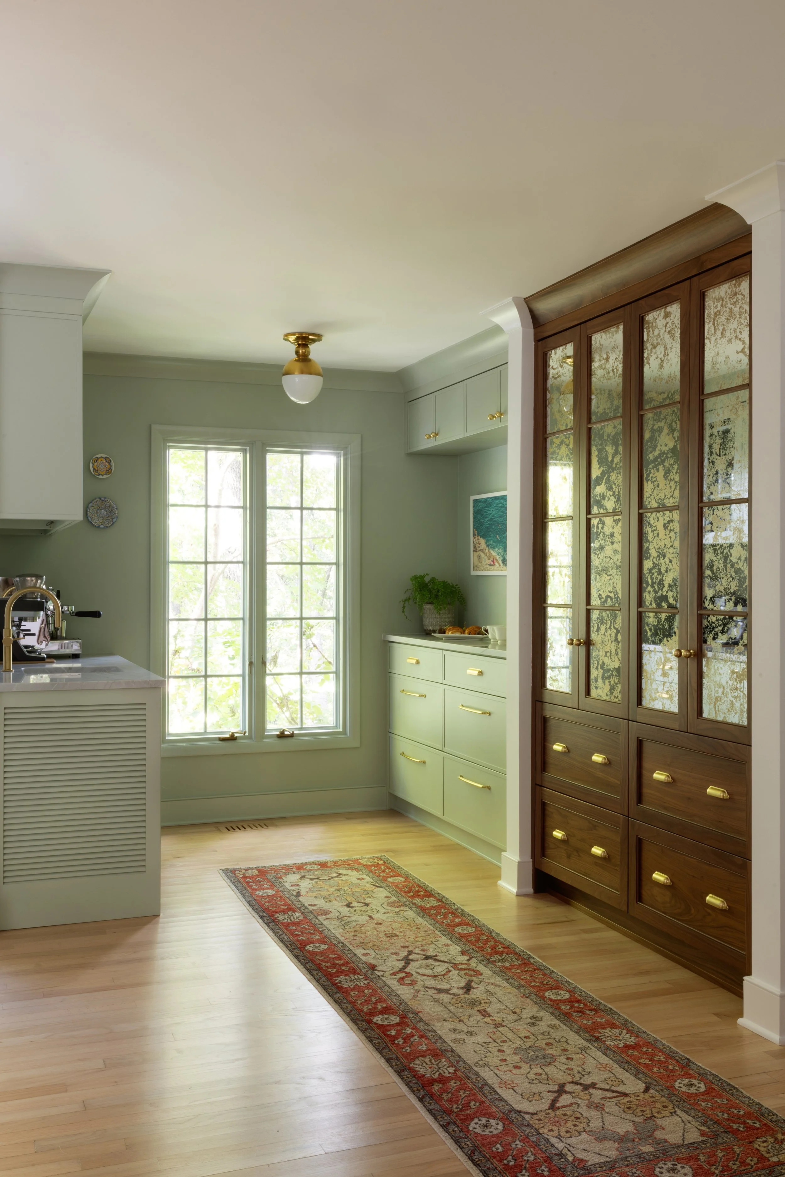 Interior of a kitchen with light green walls, a window, a wooden floor, and a rug. There is a cabinet with glass doors and gold handles, a countertop, and ceiling light fixtures.