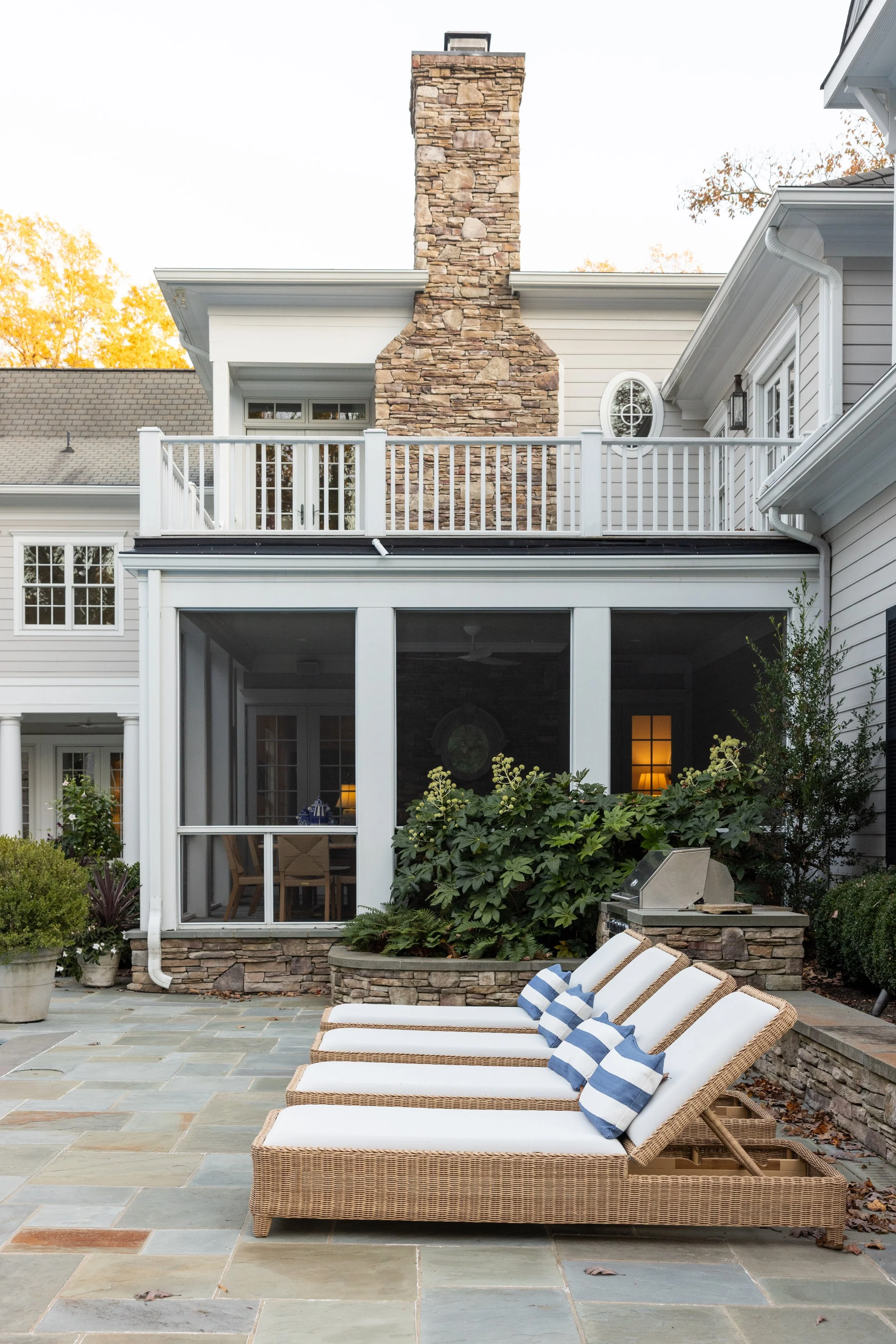 Backyard patio with wicker lounge chairs with white cushions and striped pillows, stone pavement, lush greenery, screened porch, and a two-story house with a chimney, white siding, and a balcony.