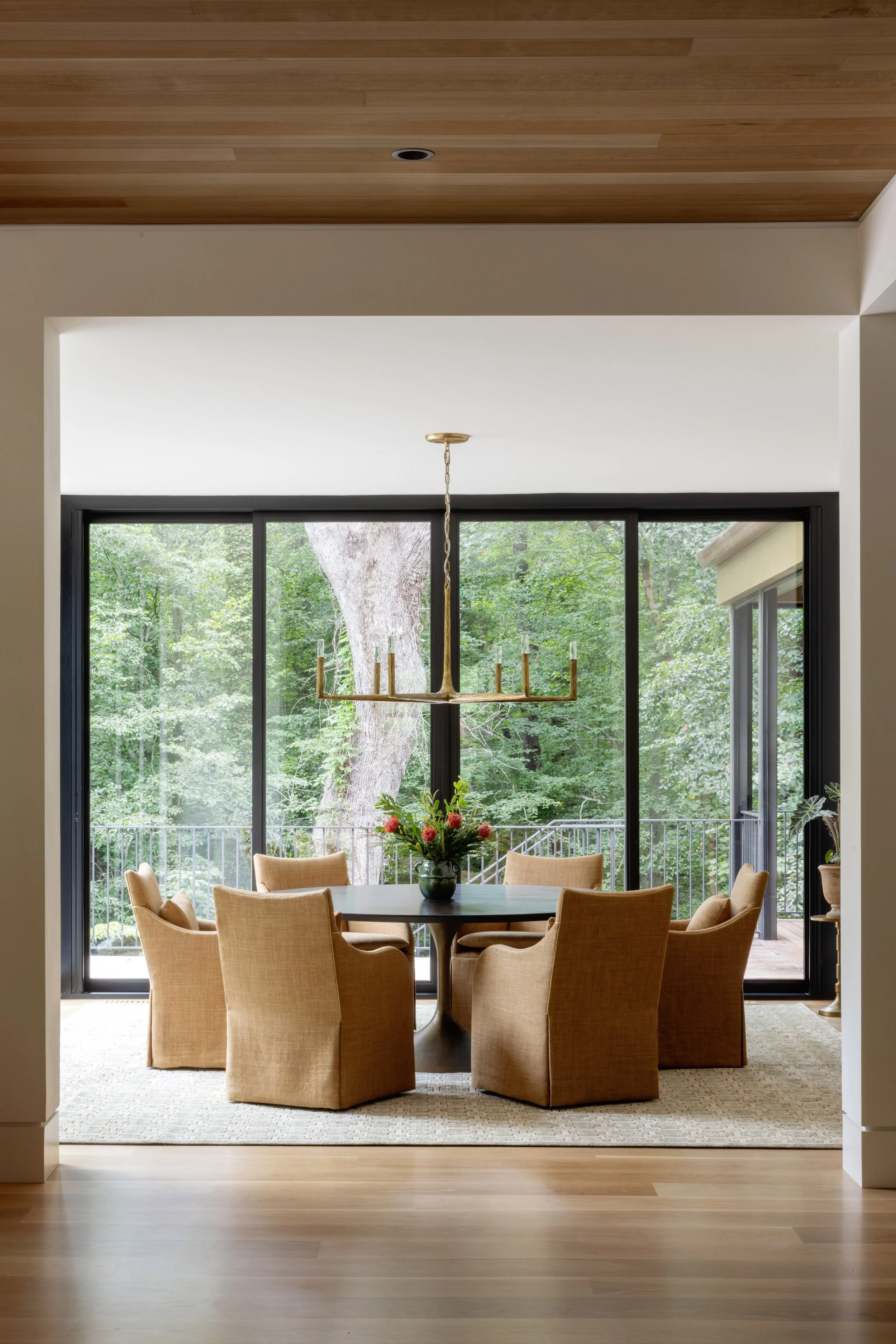 Dining room with a round table, eight beige upholstered chairs, a floral centerpiece, large windows showing greenery outside, a gold chandelier, and wooden flooring.