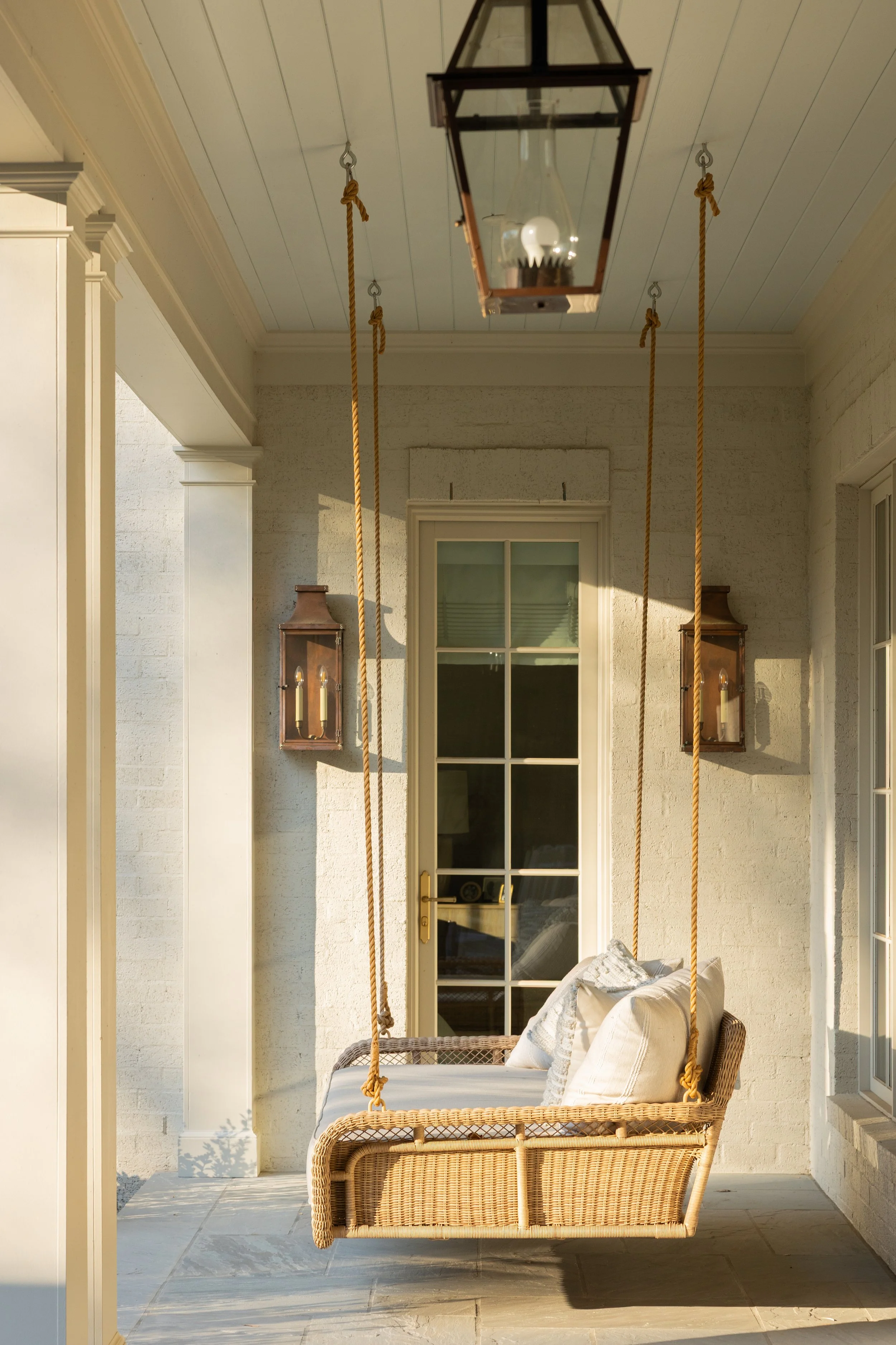 A porch area with a wicker swing bench hanging from the ceiling, decorated with pillows. The porch has stone flooring, white walls, and a glass door. Two wall-mounted lanterns are on either side of the door, and a large hanging lantern-like light fix