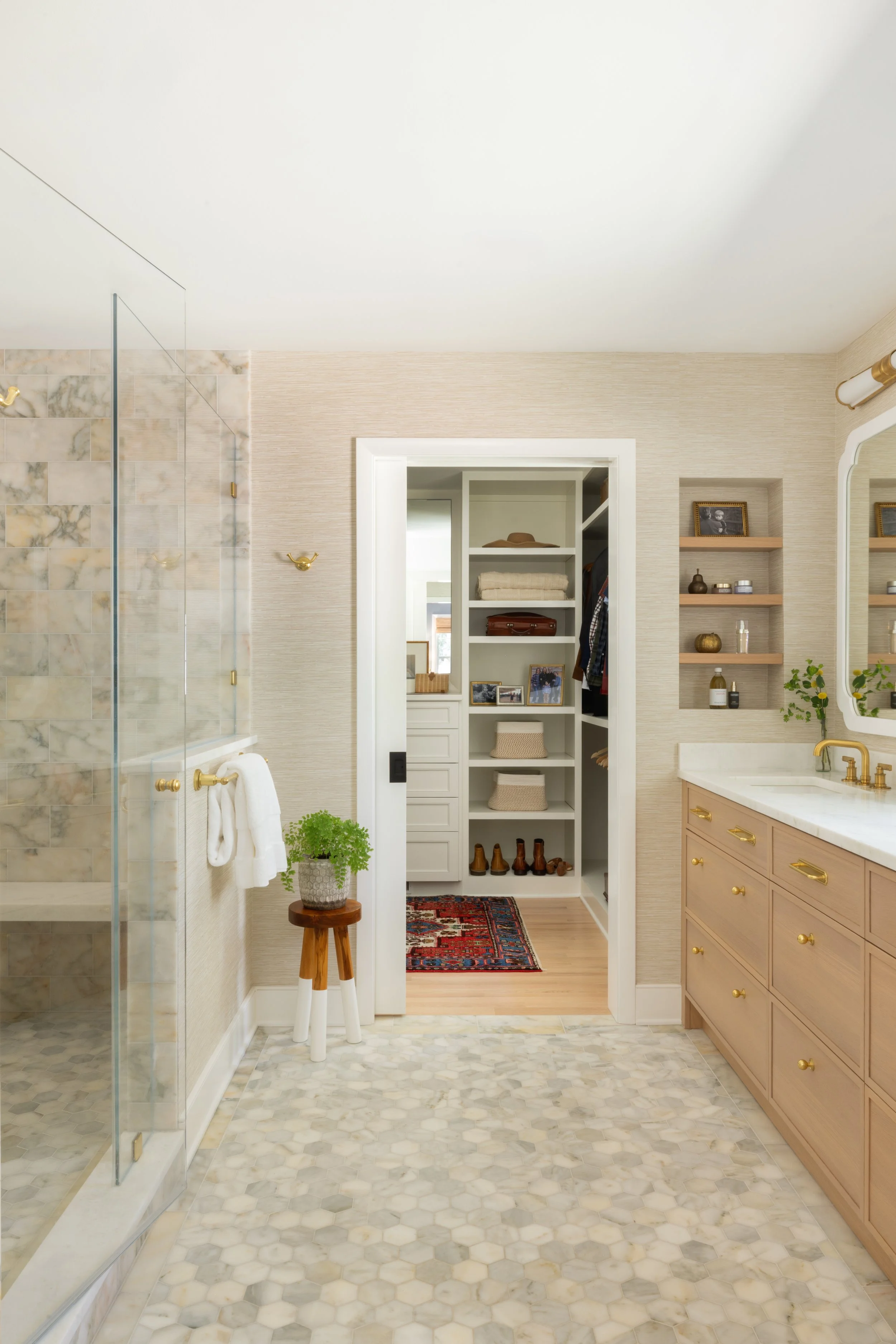Bathroom with framed mirror, light wood vanity, built-in shelves, walk-in closet with shoes, small plant on stool, and marble-tiled shower