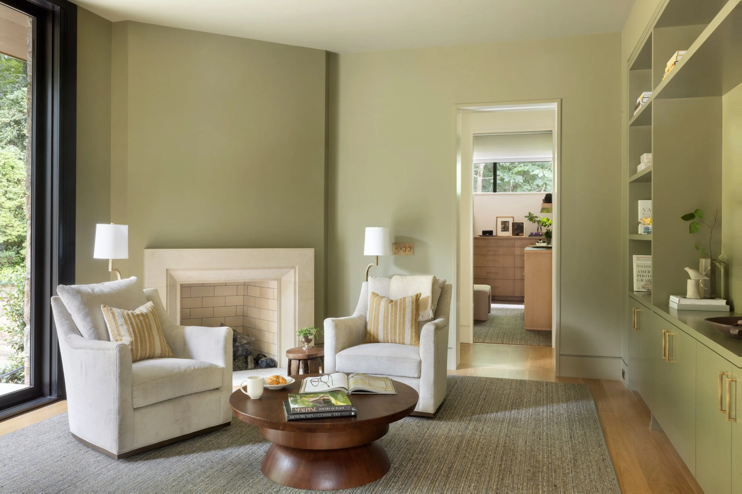 Living room with two white armchairs, a round wooden coffee table with books, a mug, and a croissant, a fireplace, large window, bookshelf, and a view into an adjacent room.