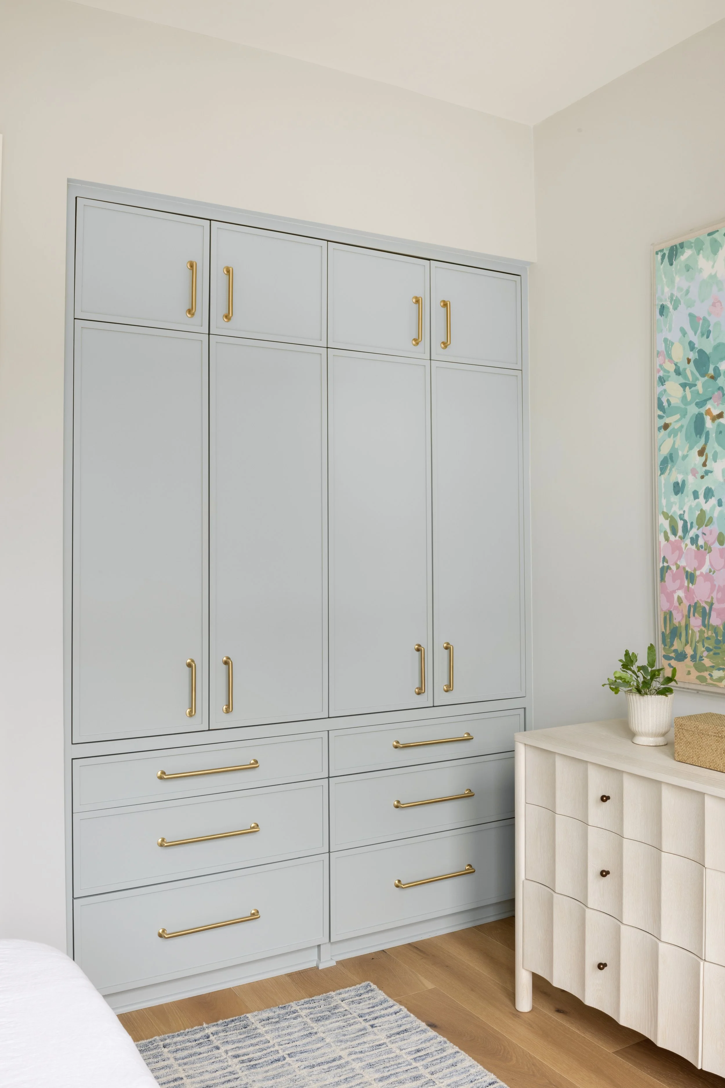 A large gray wardrobe with gold handles, situated in a bedroom corner. Next to it is a white dresser with small drawers, decorated with a small potted plant and a woven box. A colorful abstract painting hangs on the wall above the dresser.