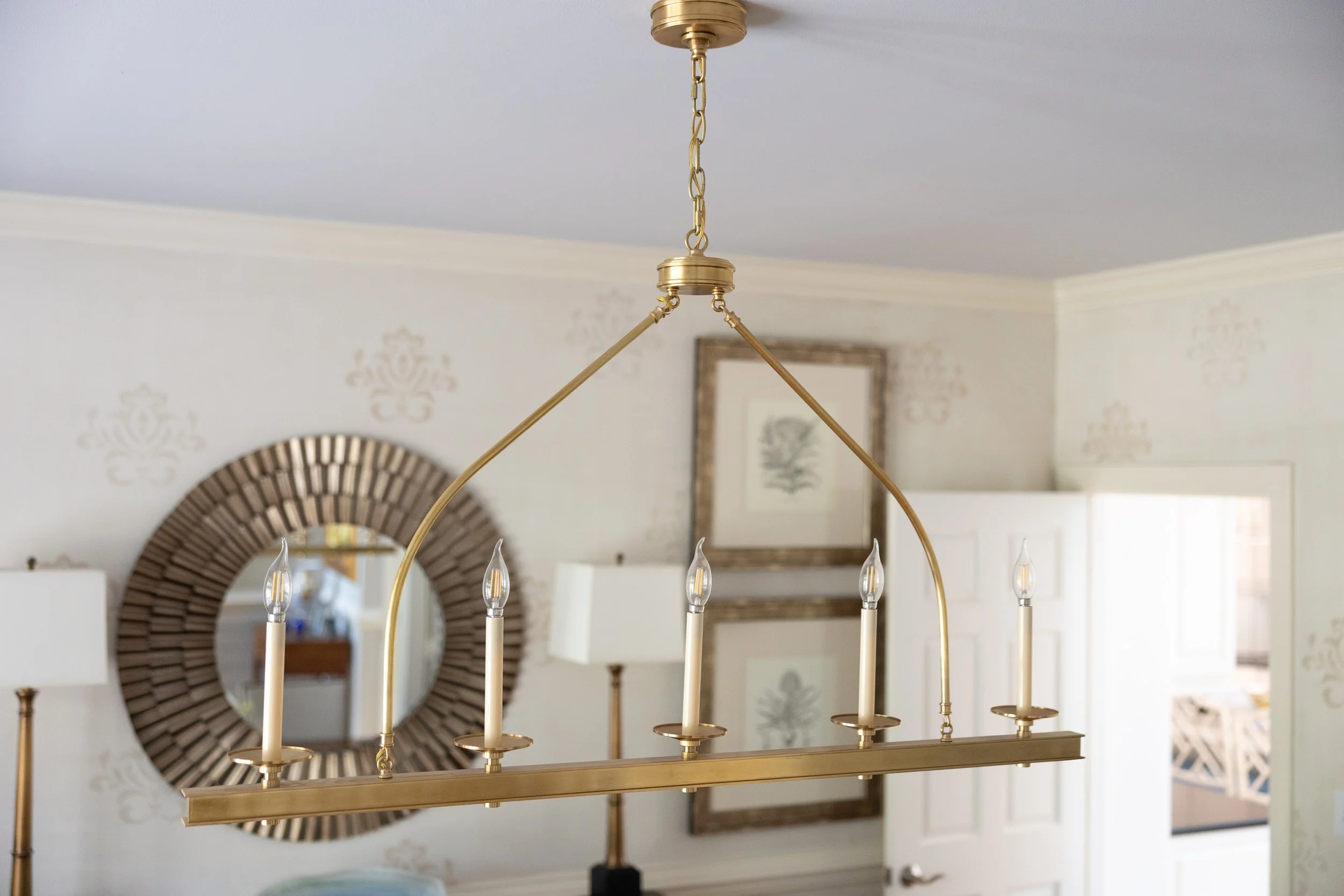 Gold chandelier with six candle-style lights hanging in a living room with framed artwork and a round mirror on the wall.