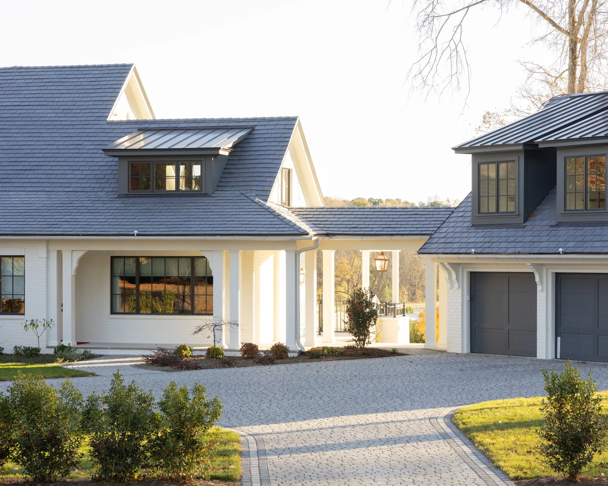 Front view of a modern house with white brick exterior, gray tiled roof, and black window frames, surrounded by a paved driveway and landscaped garden.