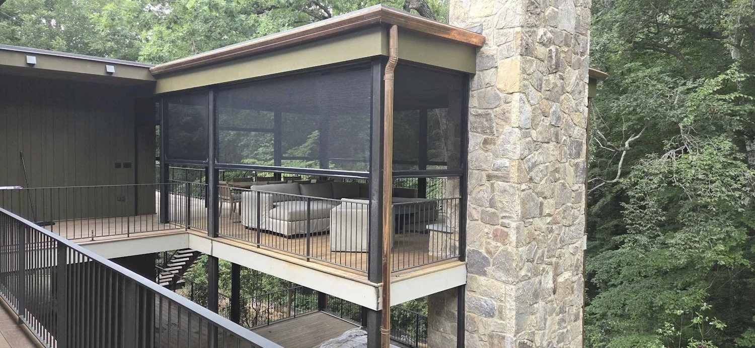 Luxury custom screened outdoor living area built by Will Johnson Building Company featuring a covered deck, black-framed screens, stone chimney structure, wood ceiling details, and seating designed for private views overlooking a wooded setting.