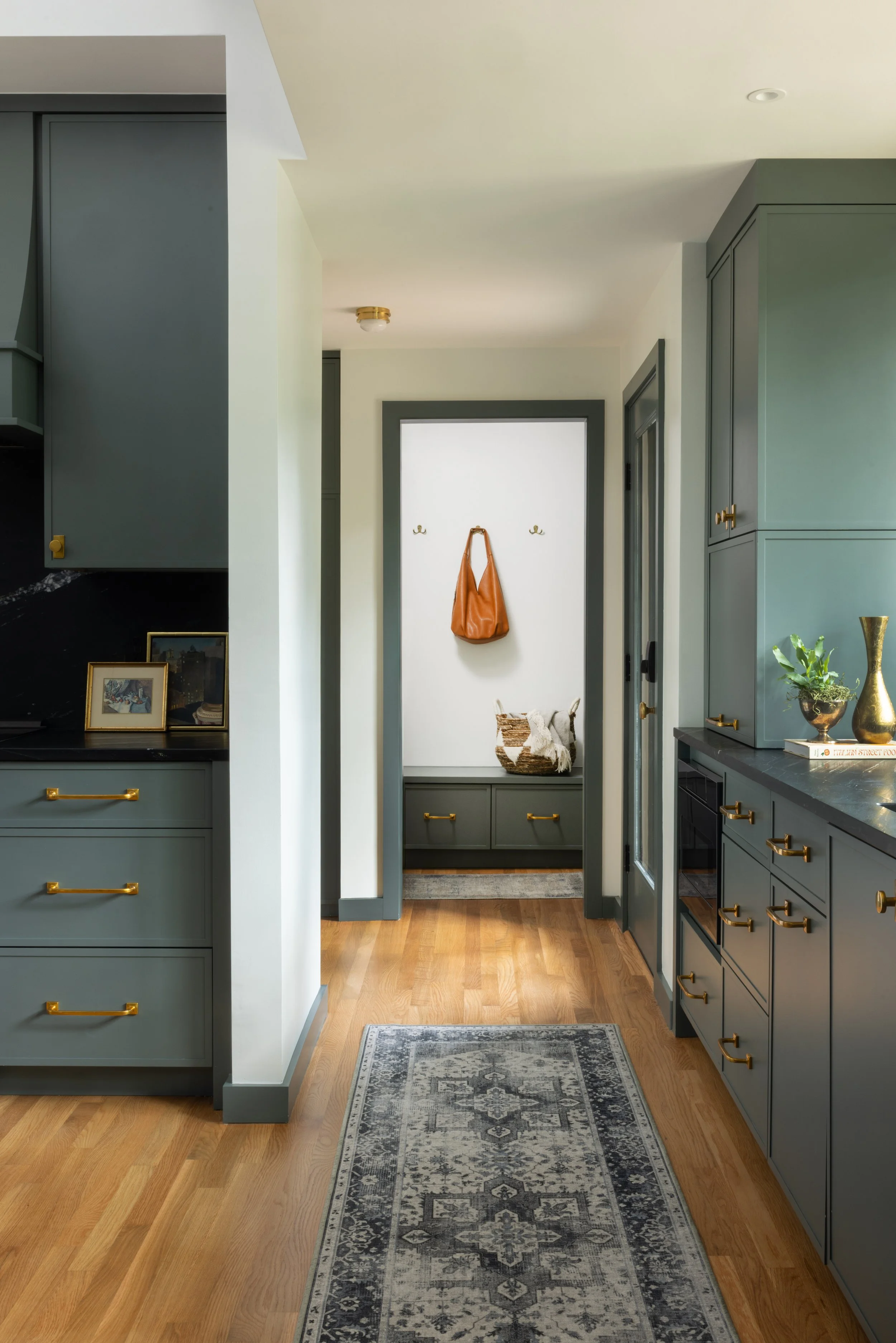 View of a modern kitchen with blue cabinets and gold hardware, a gray patterned rug on hardwood floor, and a small entryway with a white wall, hooks, and a bench with storage underneath.