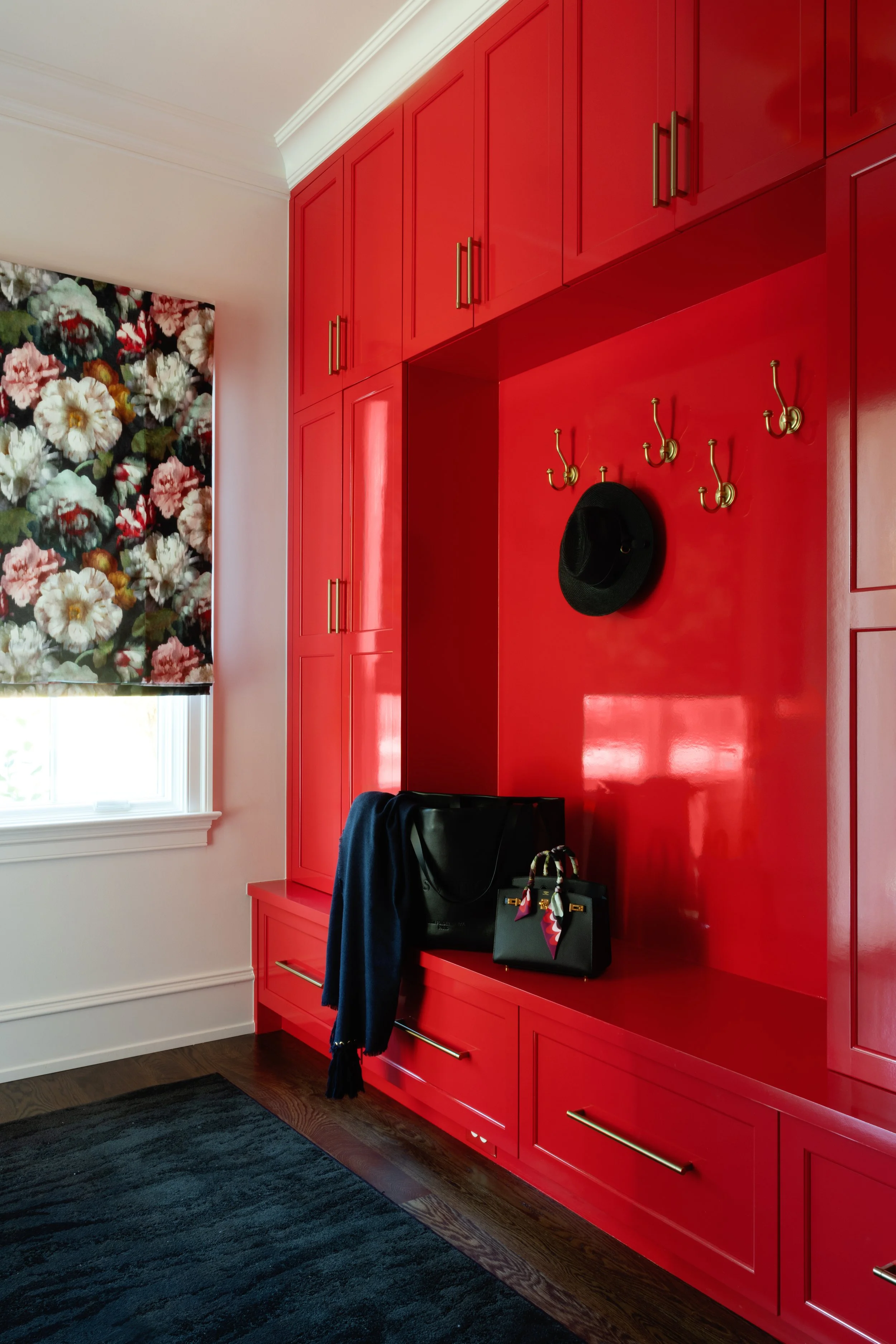 Interior of a room with bold red built-in wall cabinets, a floral window shade, black hat hanging on a gold hook, and a black bag with a scarf hanging on a red bench. Dark wooden floor and black rug.