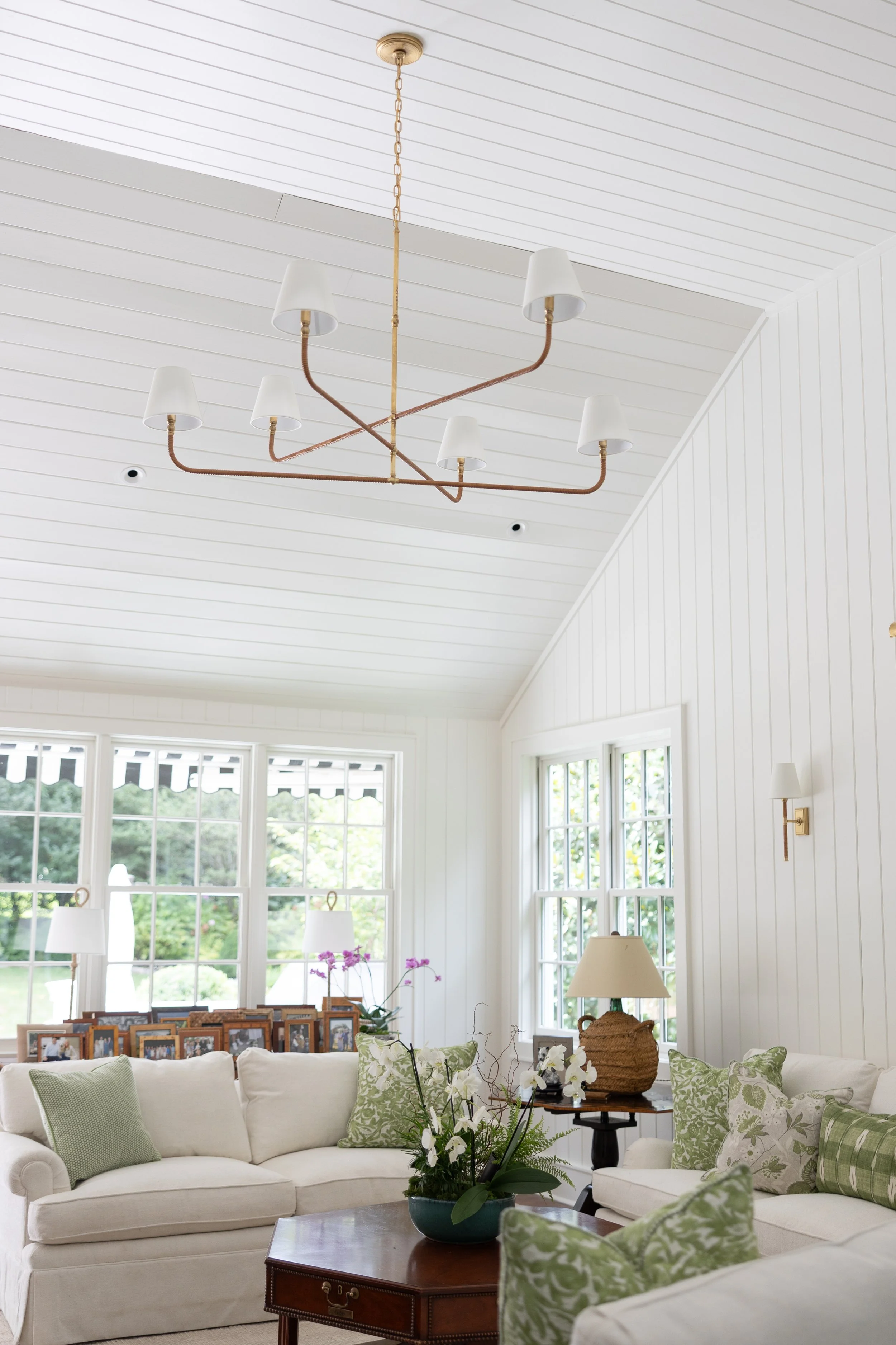 Bright living room with white walls, large windows, a modern chandelier with white shades, cream sofas with green patterned pillows, a wooden coffee table with a potted plant, and wall sconces.