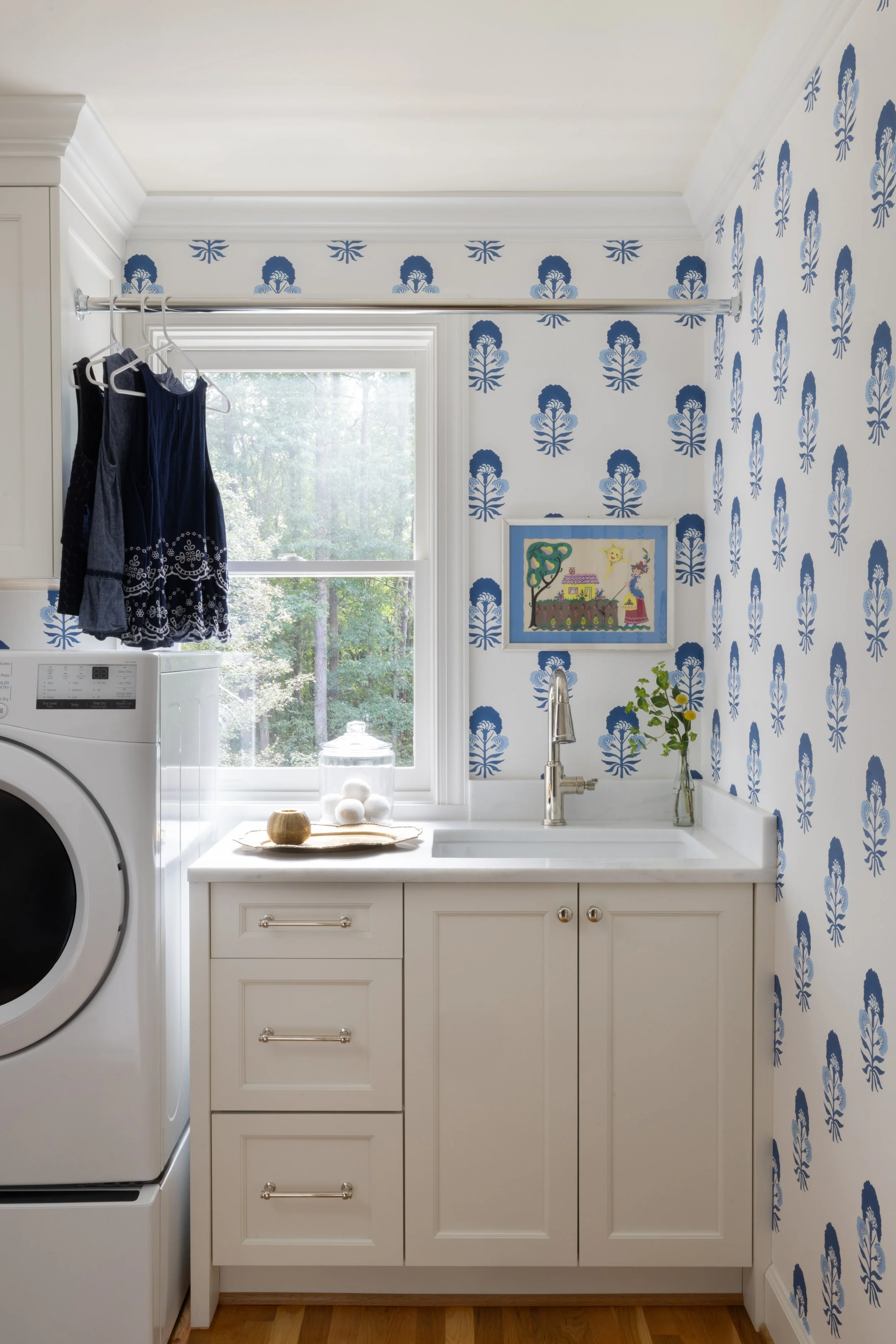 A laundry room with blue and white wallpaper, a window showing trees outside, a washing machine, a white cabinet with a countertop, a small plant in a vase, a framed drawing, and some clothes hanging on a rod.