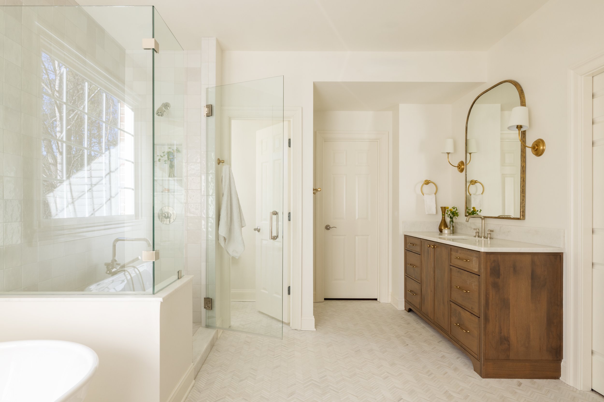 Bright bathroom with a glass-enclosed shower, a wooden vanity with a marble top, a large mirror with wall sconces, and a window reflecting trees outside.