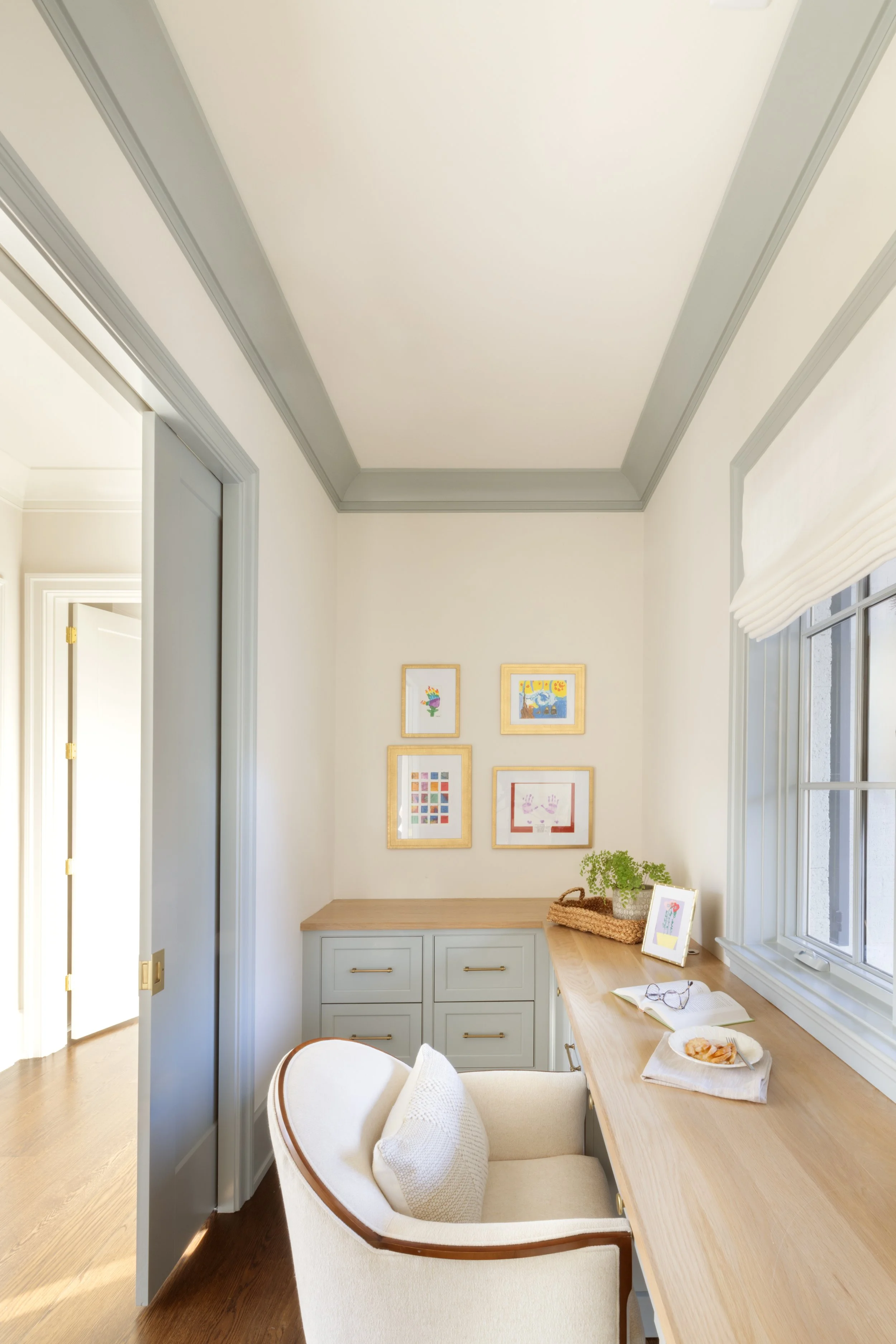 A cozy home office corner with a wooden desk, an armchair with a pillow, framed artwork on the wall, a large window with a shade, books, a potted plant, and a tray with seashells.