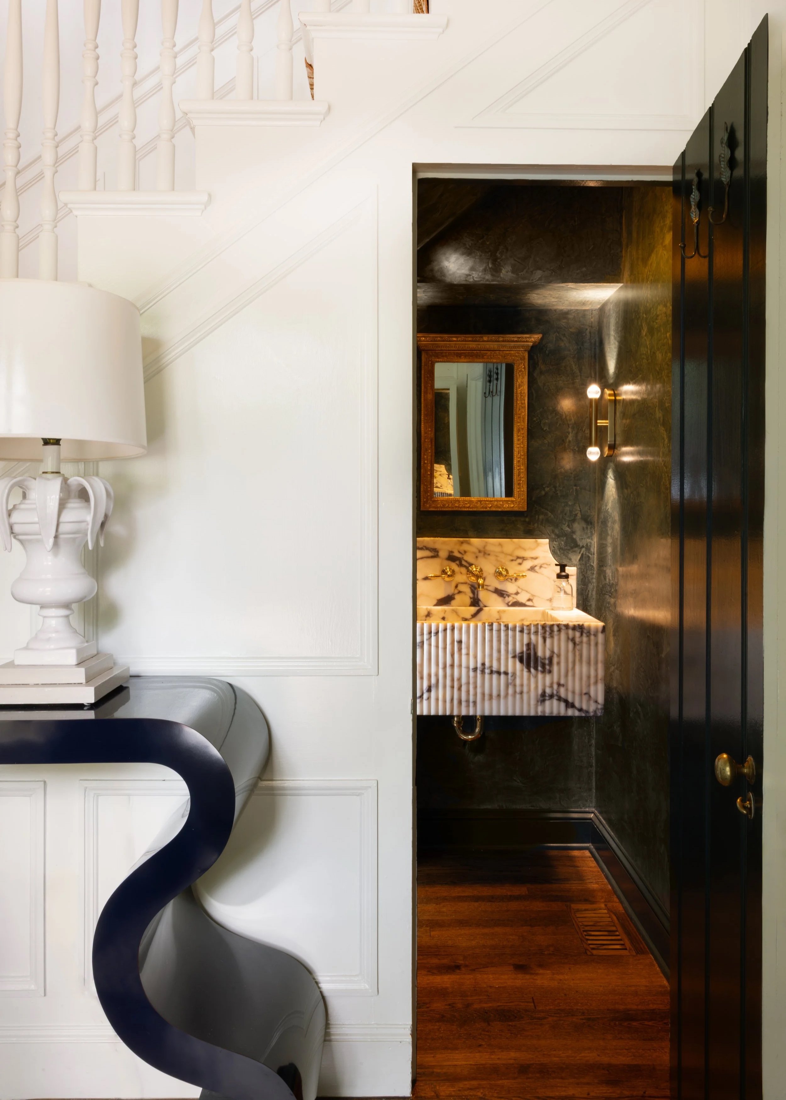 Interior view of a home's hallway showing a partial white staircase, a black door, and a glimpse into a bathroom with dark walls, a marble sink, and a mirror.,