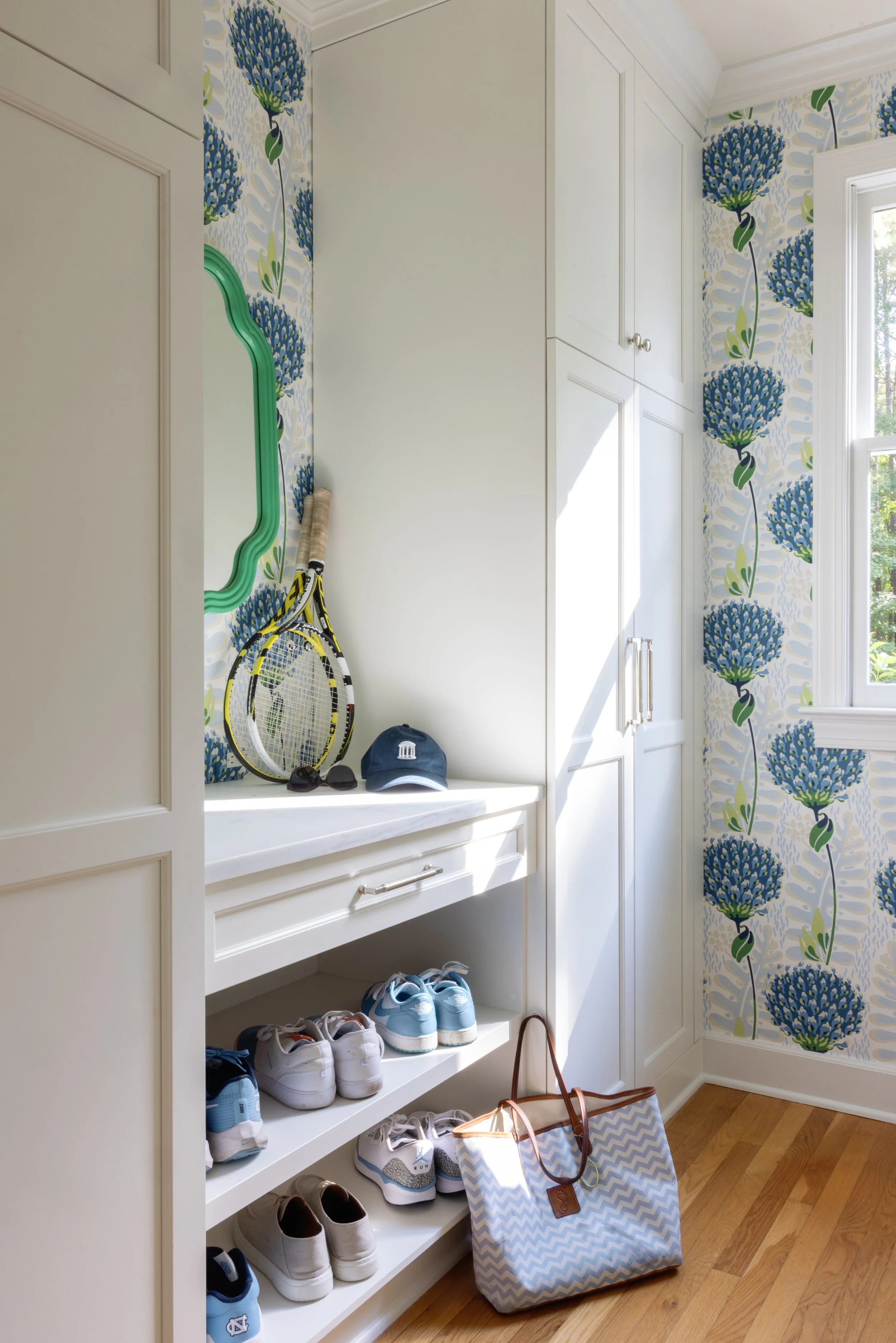 A corner of a room with white built-in cabinets, a white shoe shelf with various sneakers, a large tote bag on the floor, a tennis racket, sunglasses, and a baseball cap on a white cabinet, and blue floral wallpaper with green accents on the walls an