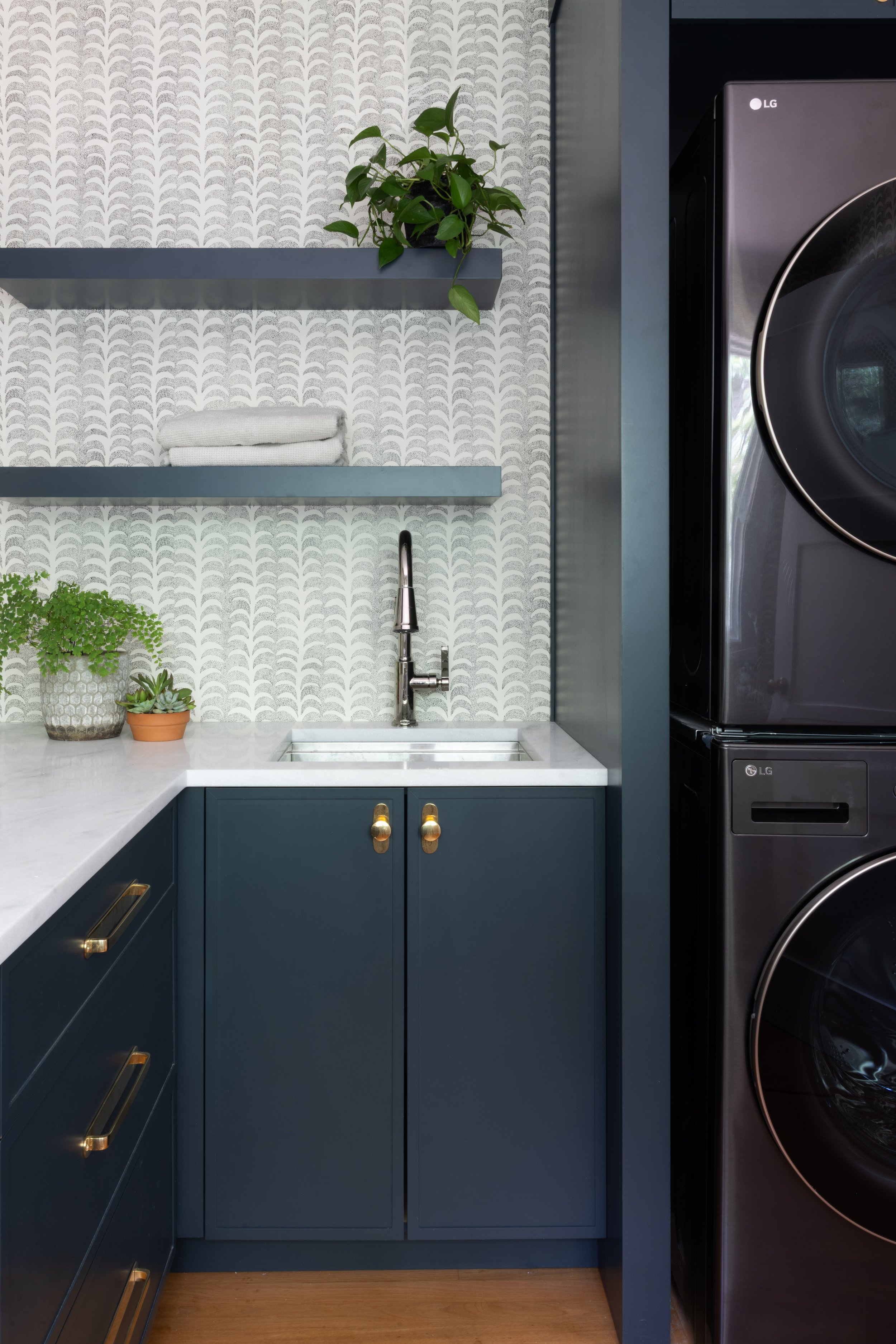 A laundry room featuring dark blue cabinets with gold handles, a white marble countertop, a small white sink with a chrome faucet, decorative plants in pots, open gray shelves with stacked towels and a plant, patterned gray and white wallpaper, and a