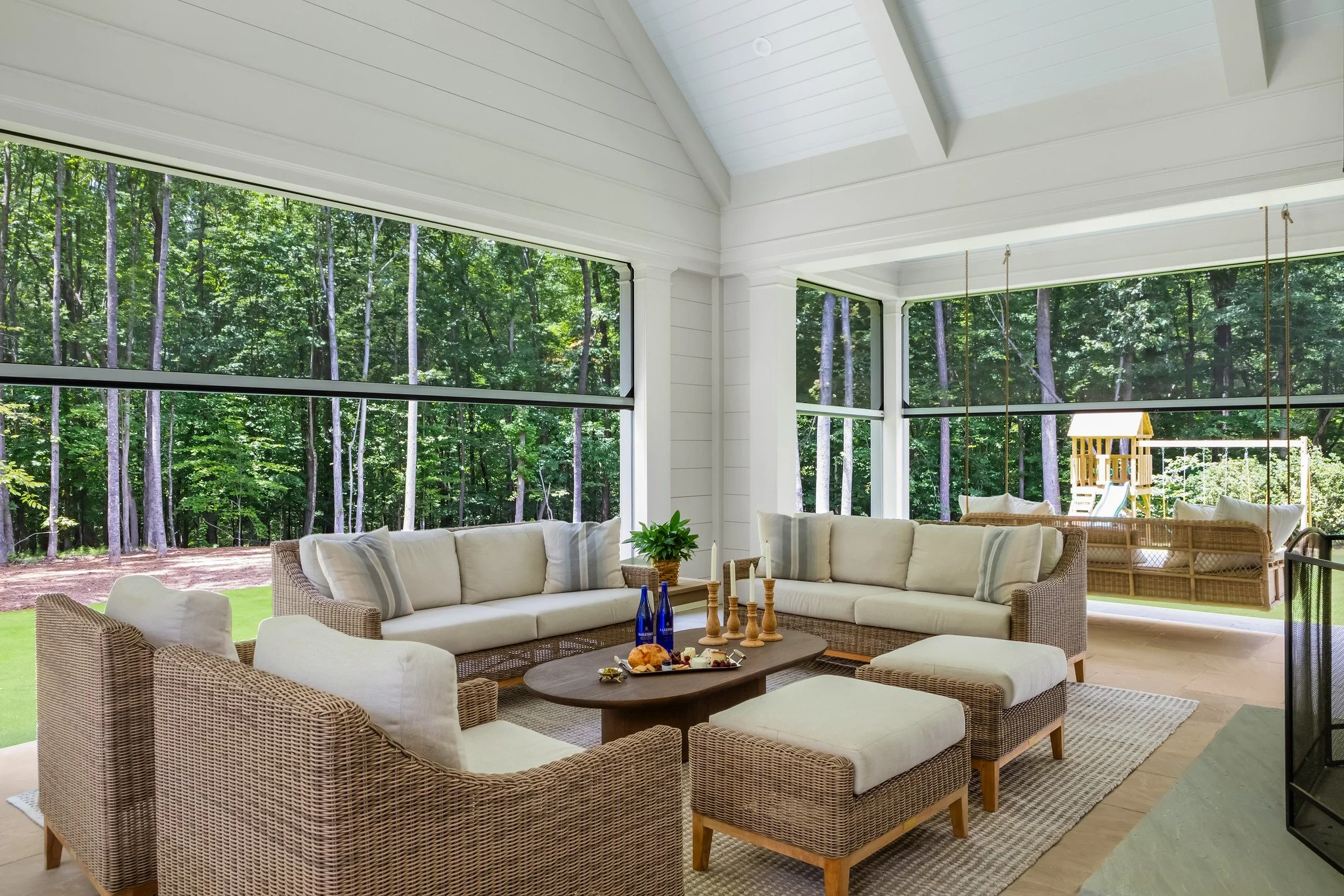Bright sunroom with large windows overlooking a wooded backyard, wicker sofa and armchairs with cream cushions, a wooden coffee table with candles and bottles, and a children's playset outside.
