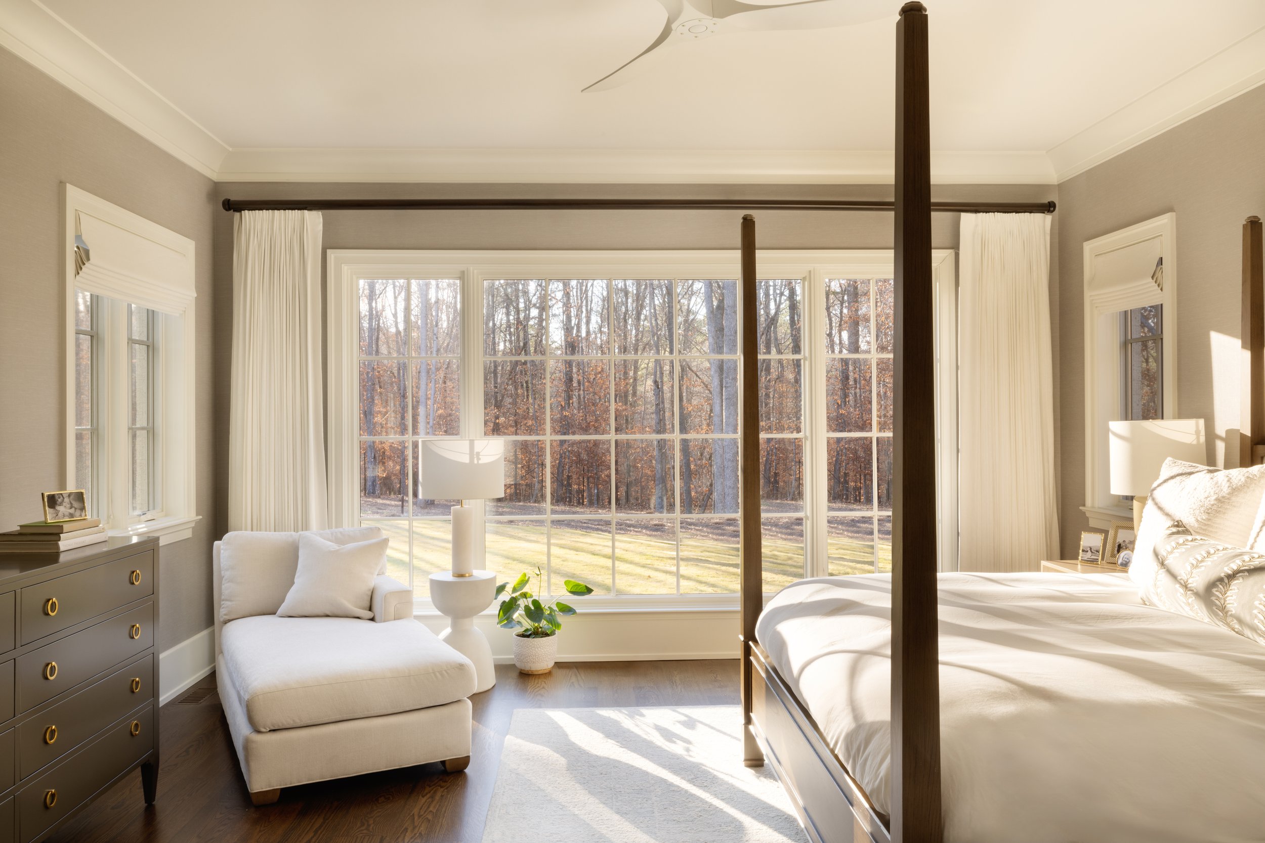 A bright bedroom with large windows overlooking a wooded outdoor area. The room features a four-poster bed with white bedding, a cream-colored chaise lounge with pillows, a gray dresser with gold drawer pulls, and a white table lamp. A potted plant i
