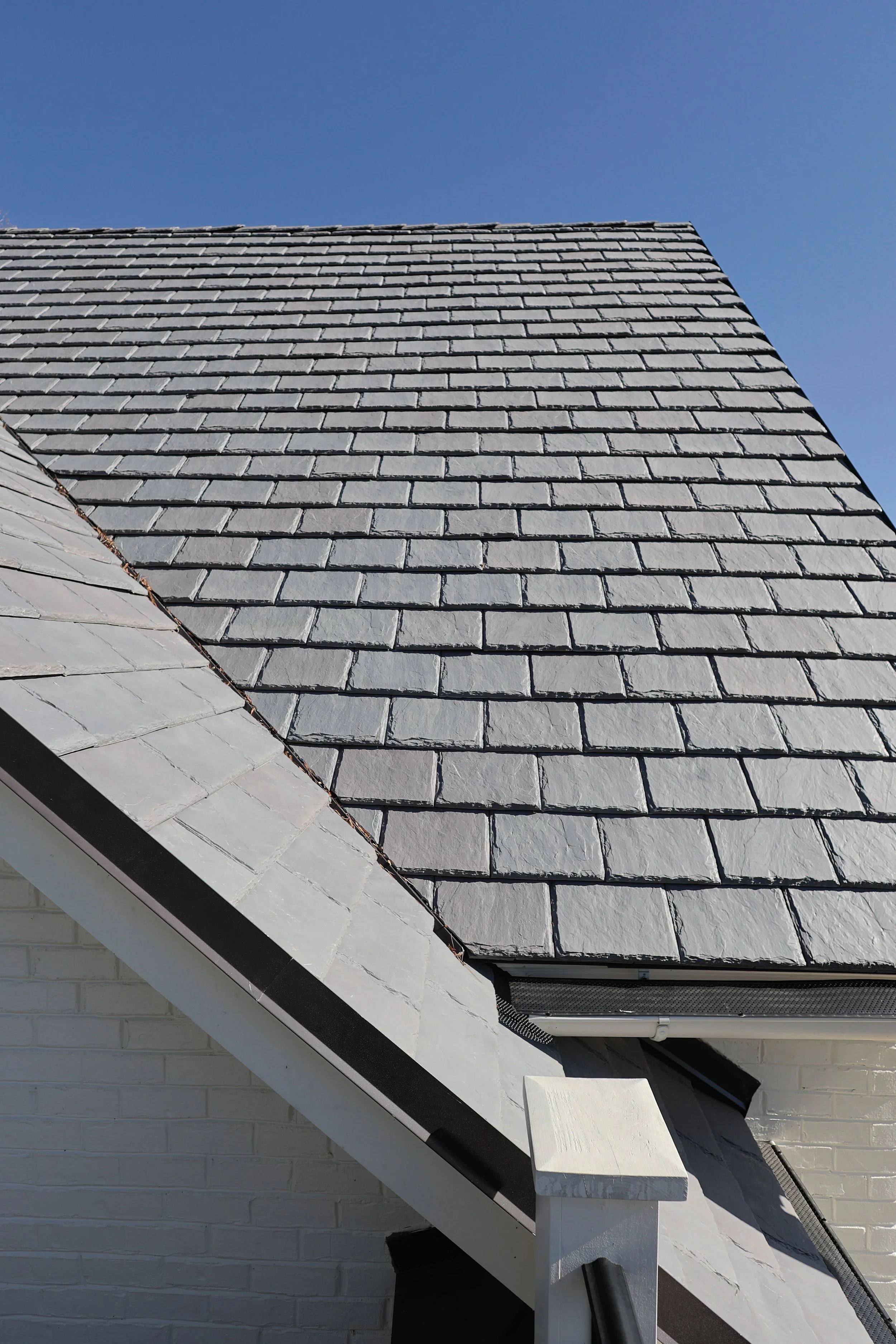 Close-up of a house roof with dark gray shingles and a section of white painted brick wall under a clear blue sky.