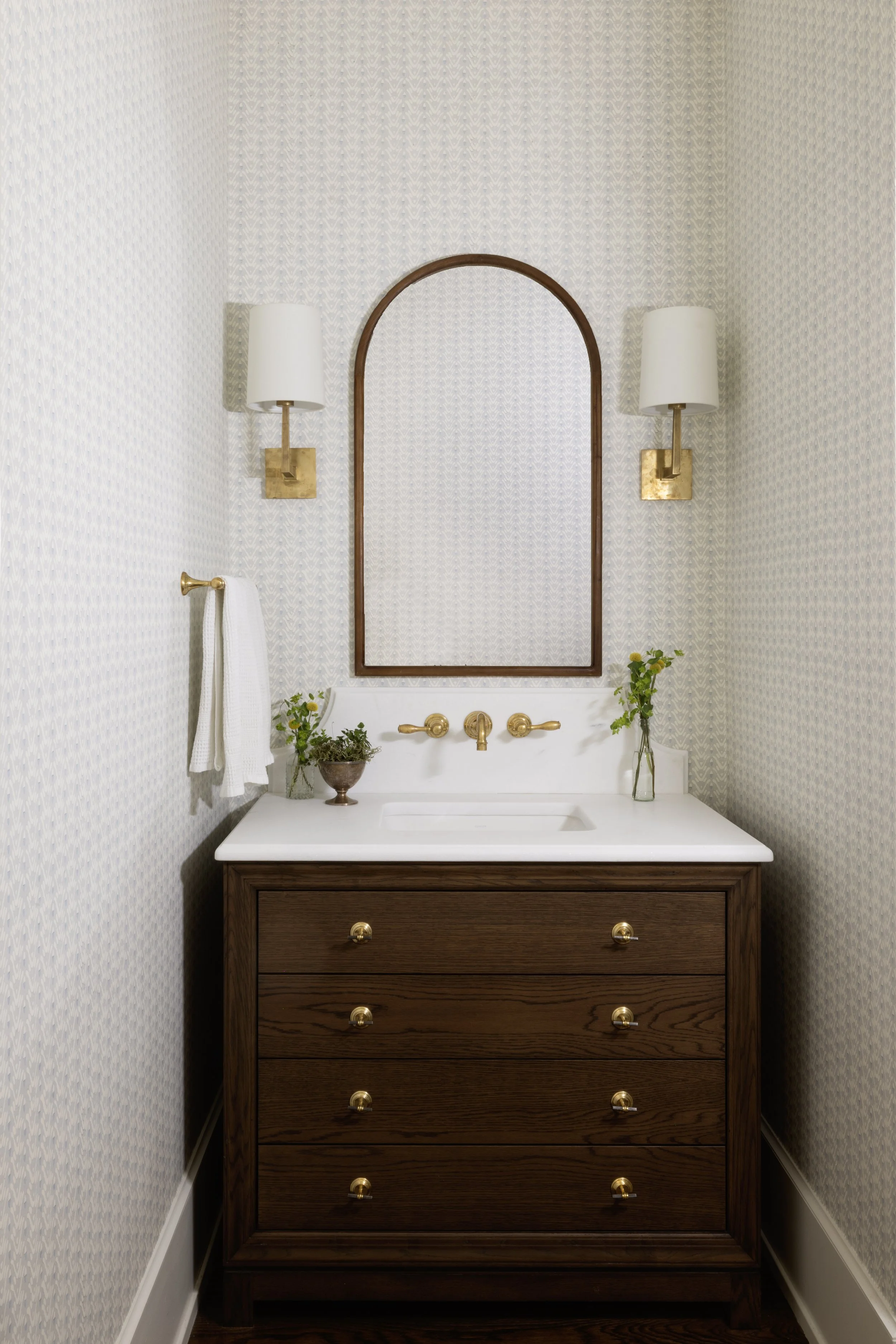 A bathroom vanity with a white countertop and a brown wooden cabinet. Above the sink, there's a large mirror with an arched wooden frame. On each side of the mirror, there are wall-mounted light fixtures with white shades and gold bases. On the left 