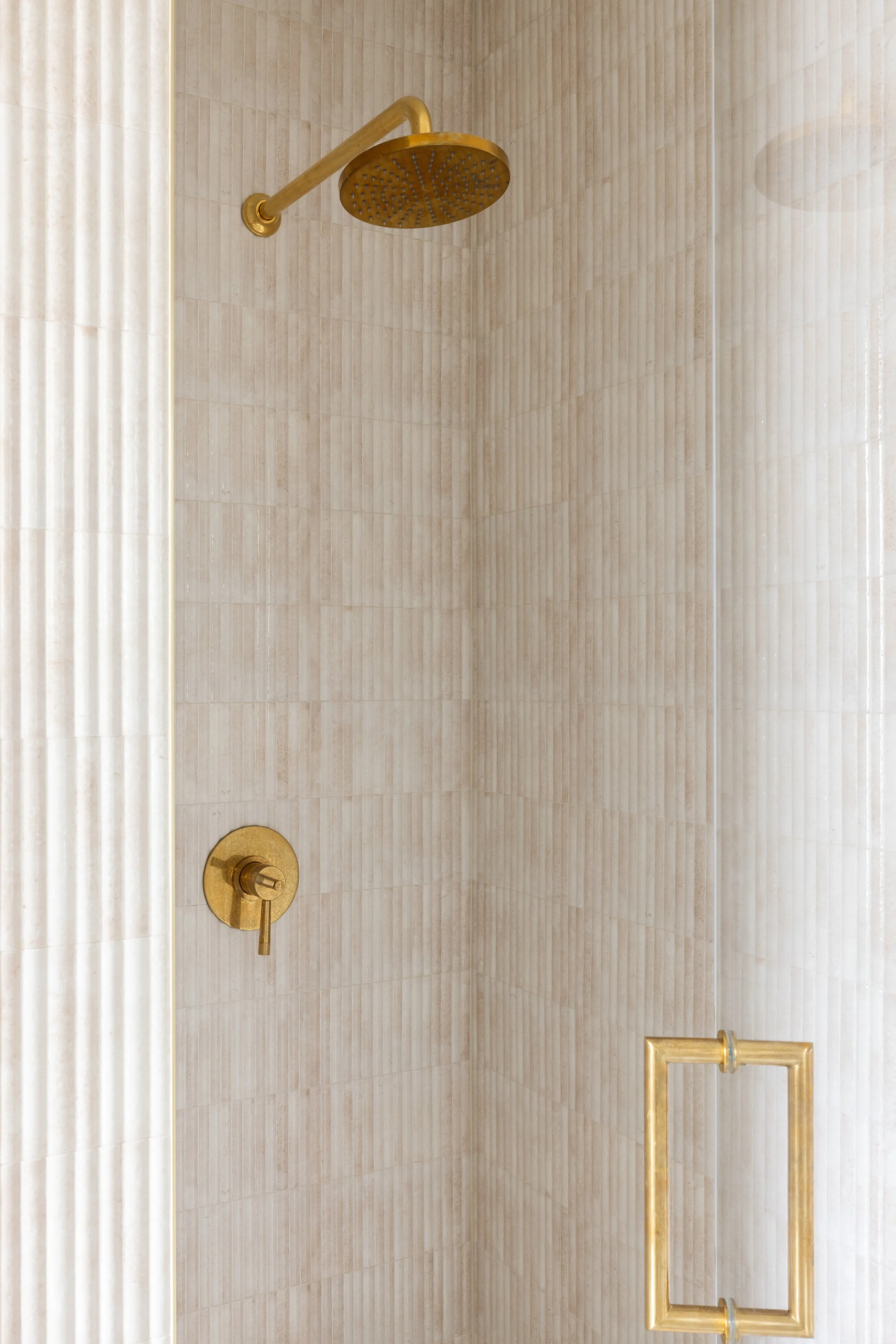 A shower with beige ceramic tiles, a gold rain showerhead, a gold control handle, and a gold towel bar.