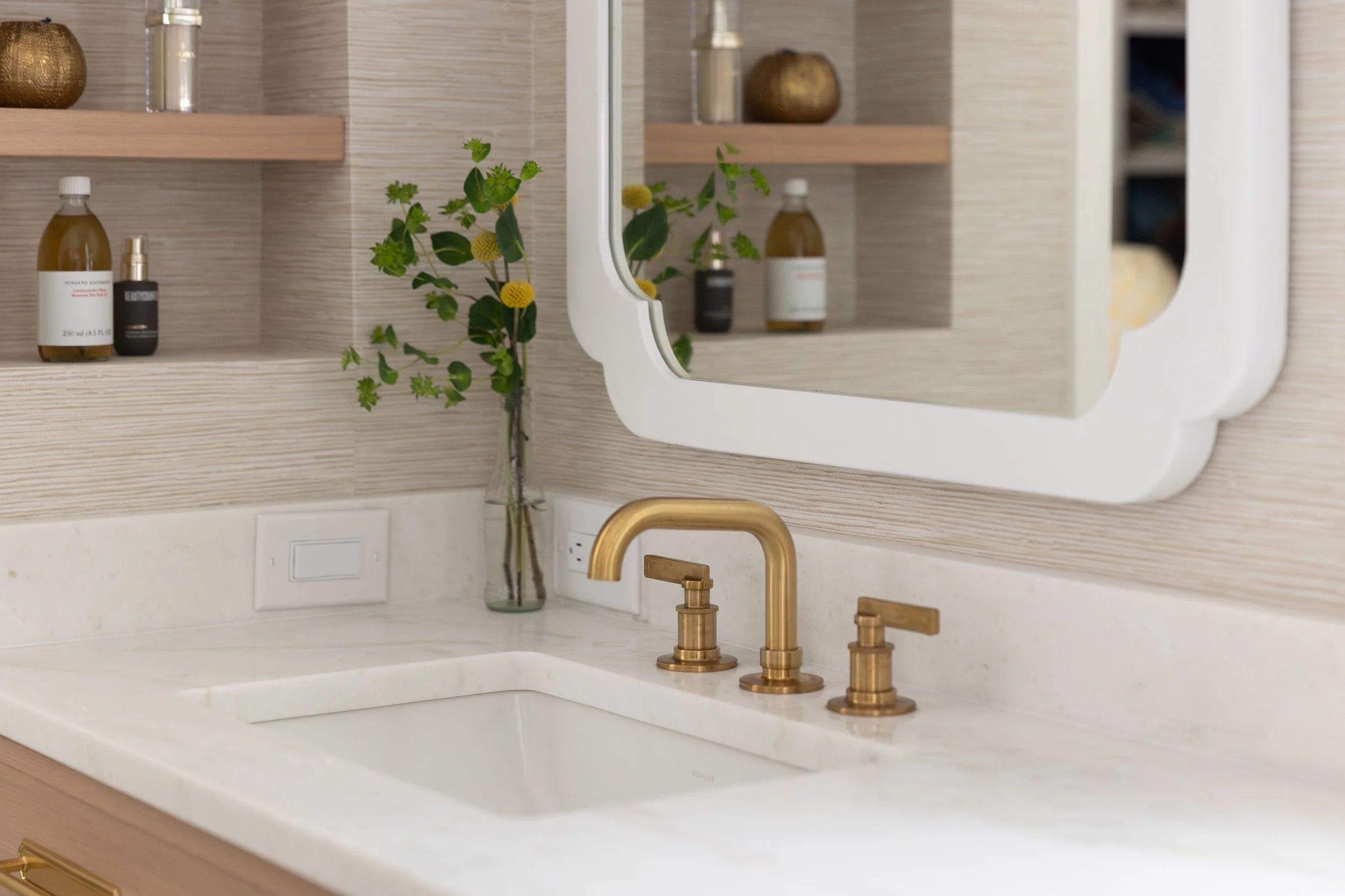 Bathroom sink with gold fixtures, a white countertop, a rectangular undermount sink, a mirror with a white scalloped border, a glass vase with greenery, and skincare bottles on a shelf.