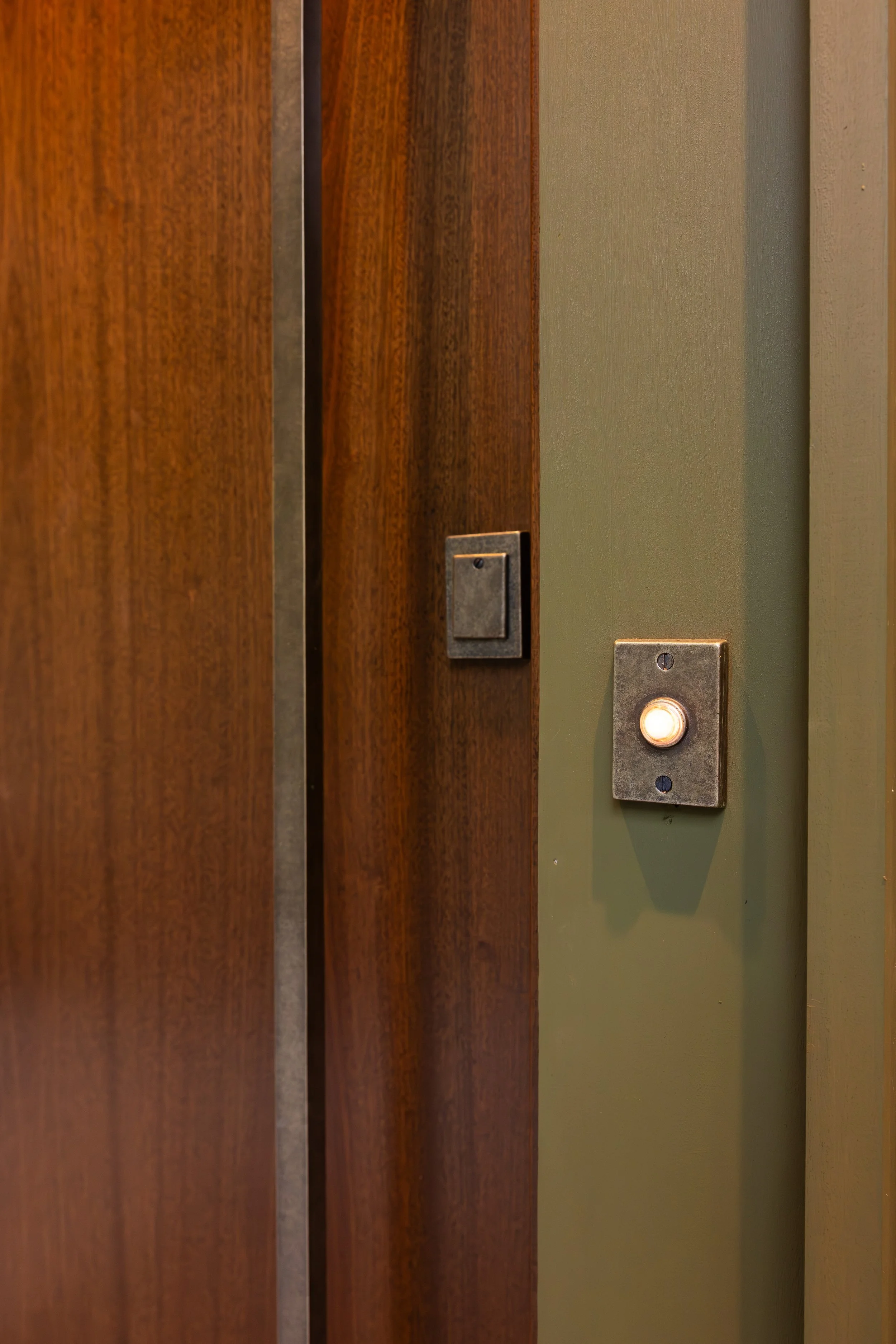 Close-up of a wooden elevator door, an illuminated call button on a green wall, and a metallic elevator switch box.