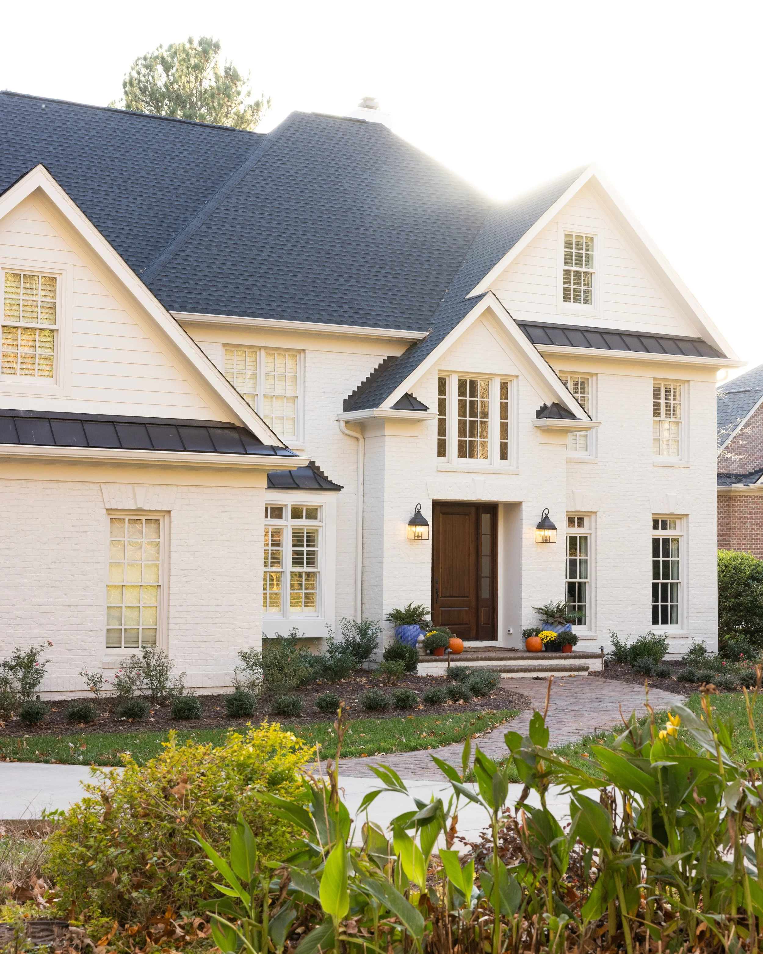 A large white house with a black roof and a wooden front door decorated with pumpkins and potted plants, surrounded by a landscaped yard with bushes and a path leading to the entrance, under a sunny sky.