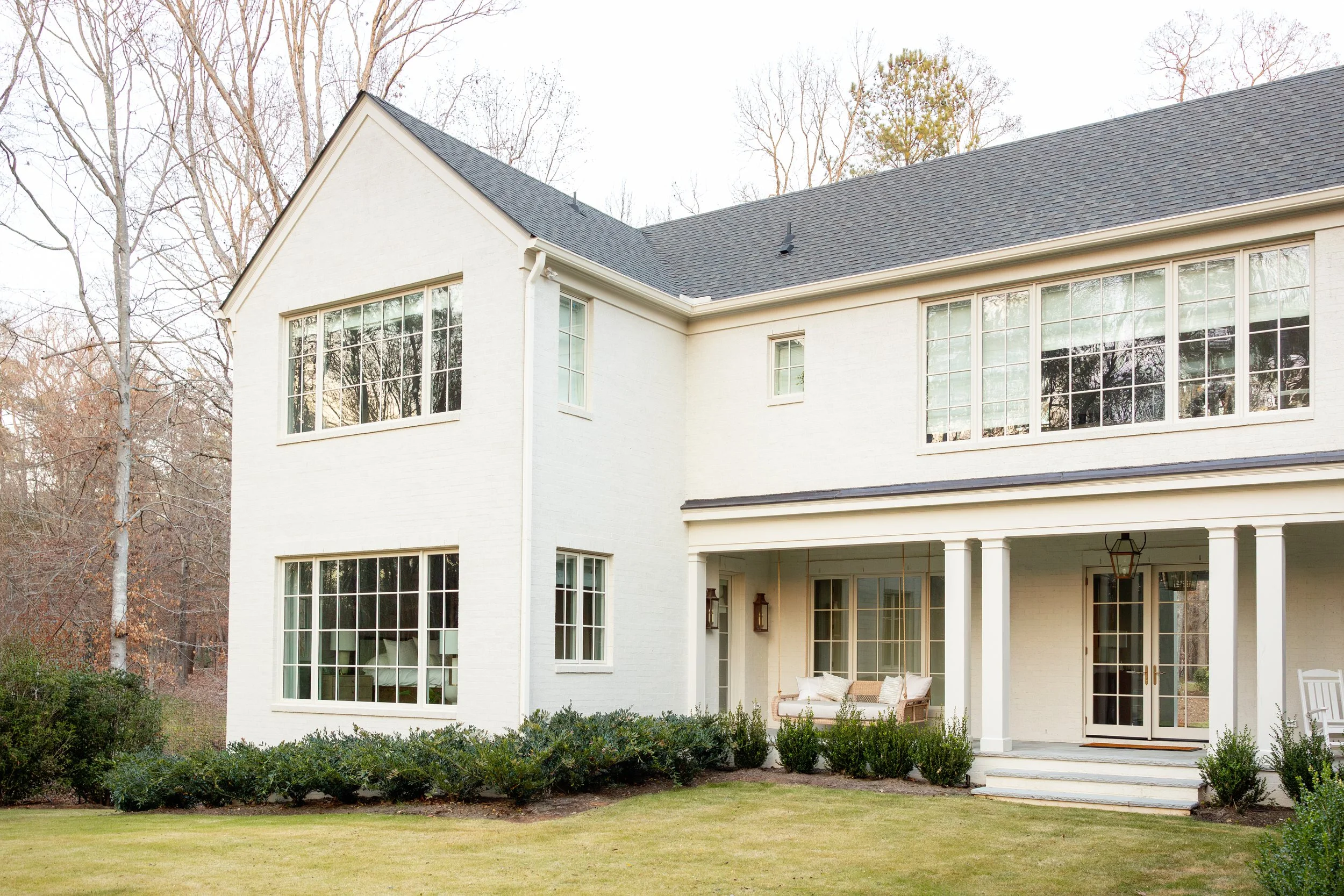 A white two-story house with large windows, a porch with outdoor furniture, surrounded by greenery and trees.