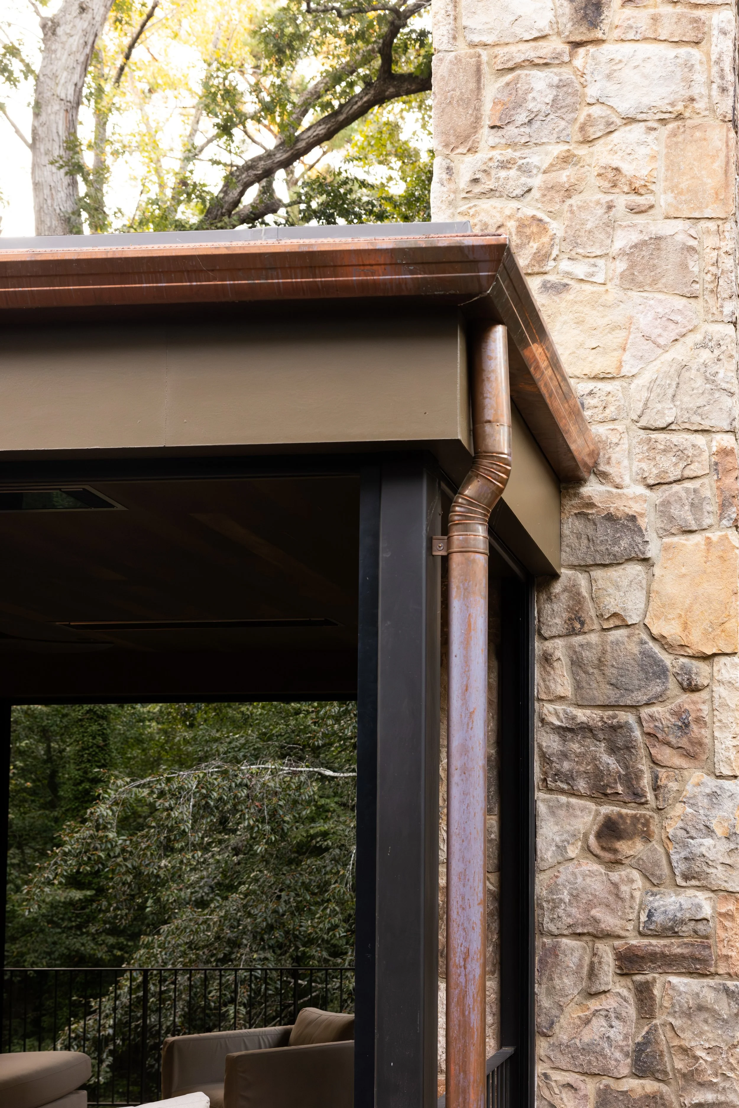 Close-up of a corner of a building featuring a stone wall, black metal support beam, and a rusted copper downspout.