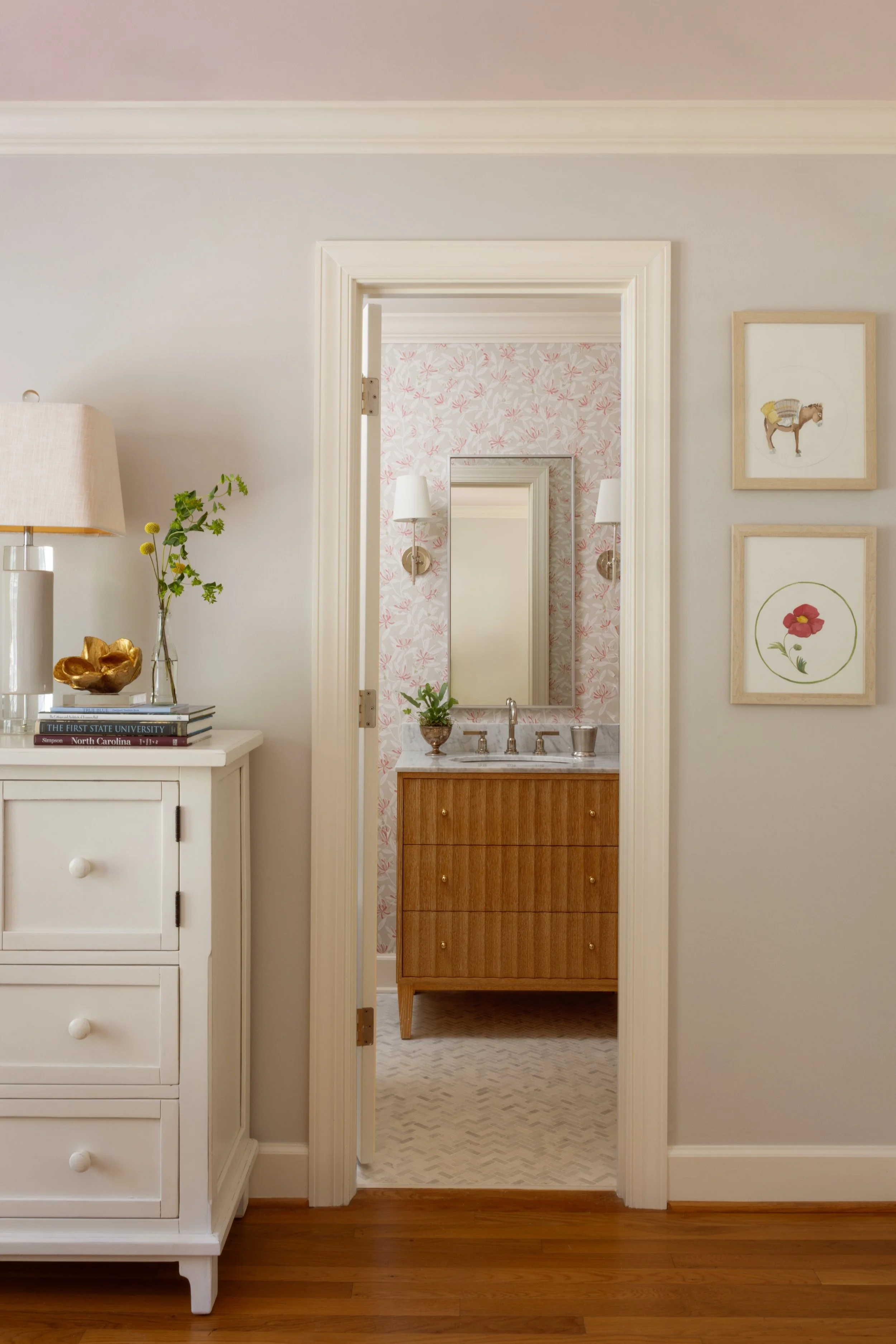 A view through a doorway into a bathroom with pink floral wallpaper, a wooden vanity with a mirror, and two wall sconces.