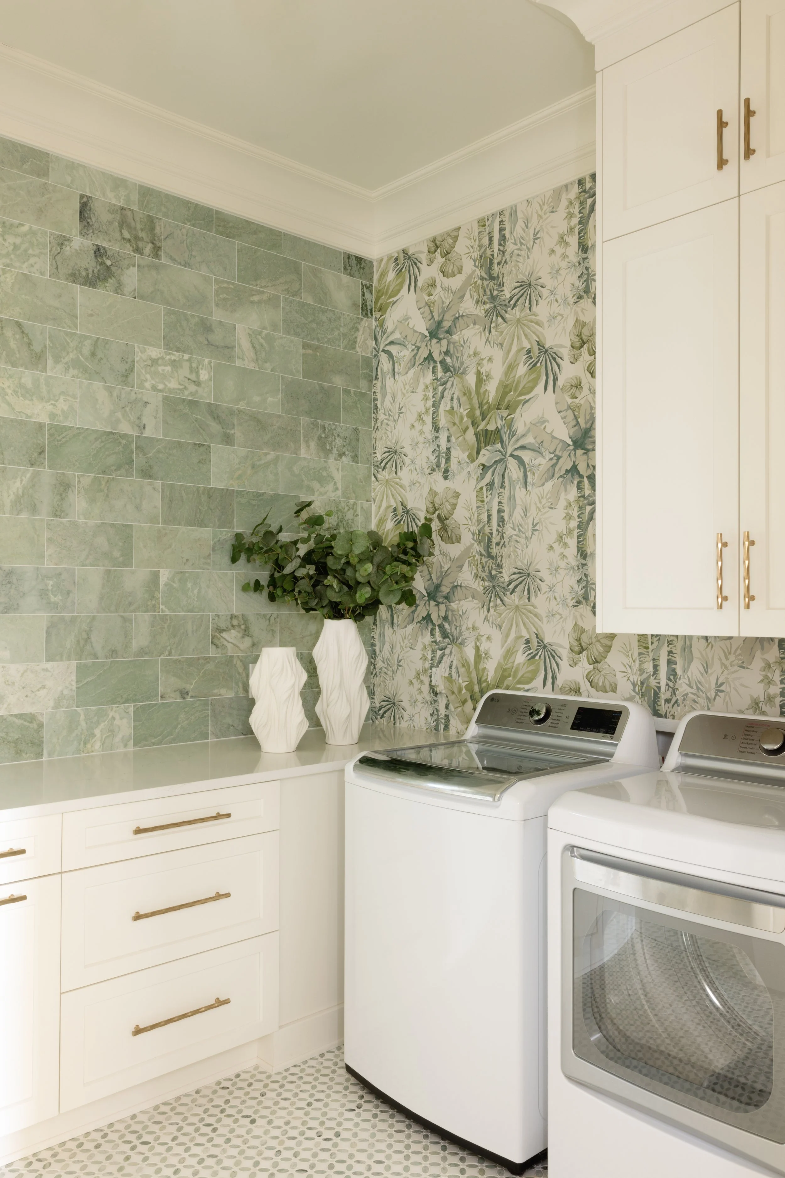 Laundry room with white washer and dryer, white cabinets, green tile and floral wallpaper, and two white vases with greenery.