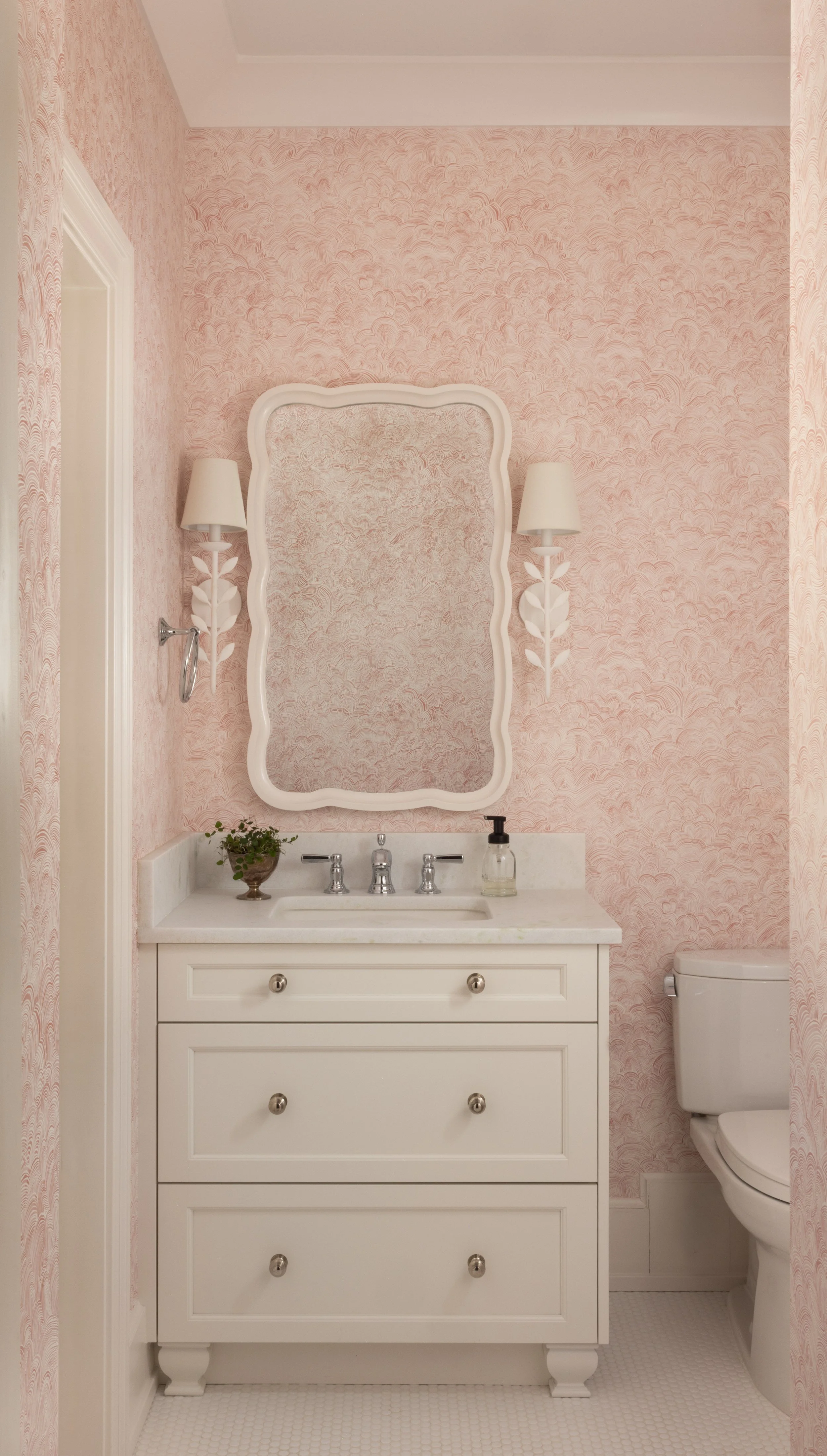 A white bathroom vanity with three drawers, a marble countertop, and a silver faucet. A small potted plant and a soap dispenser sit on the countertop. Above is a wavy-edged mirror, flanked by two wall-mounted lamps with white shades. To the right is 