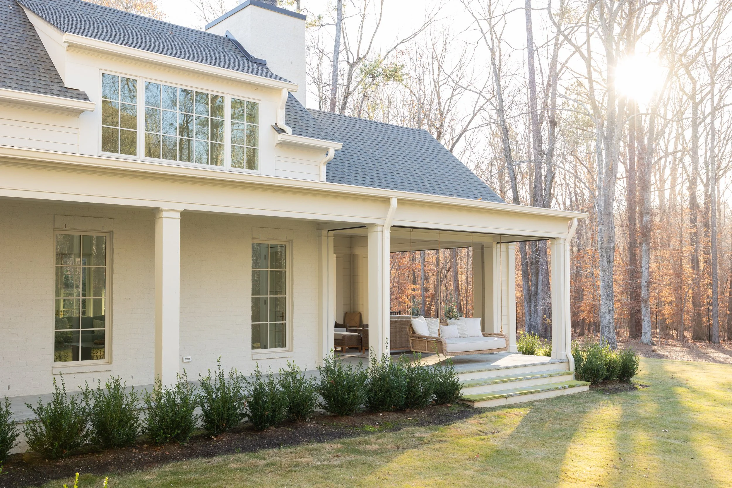 The image shows the back of a white house with a covered porch, outdoor seating, and a well-maintained lawn with trees in the background.