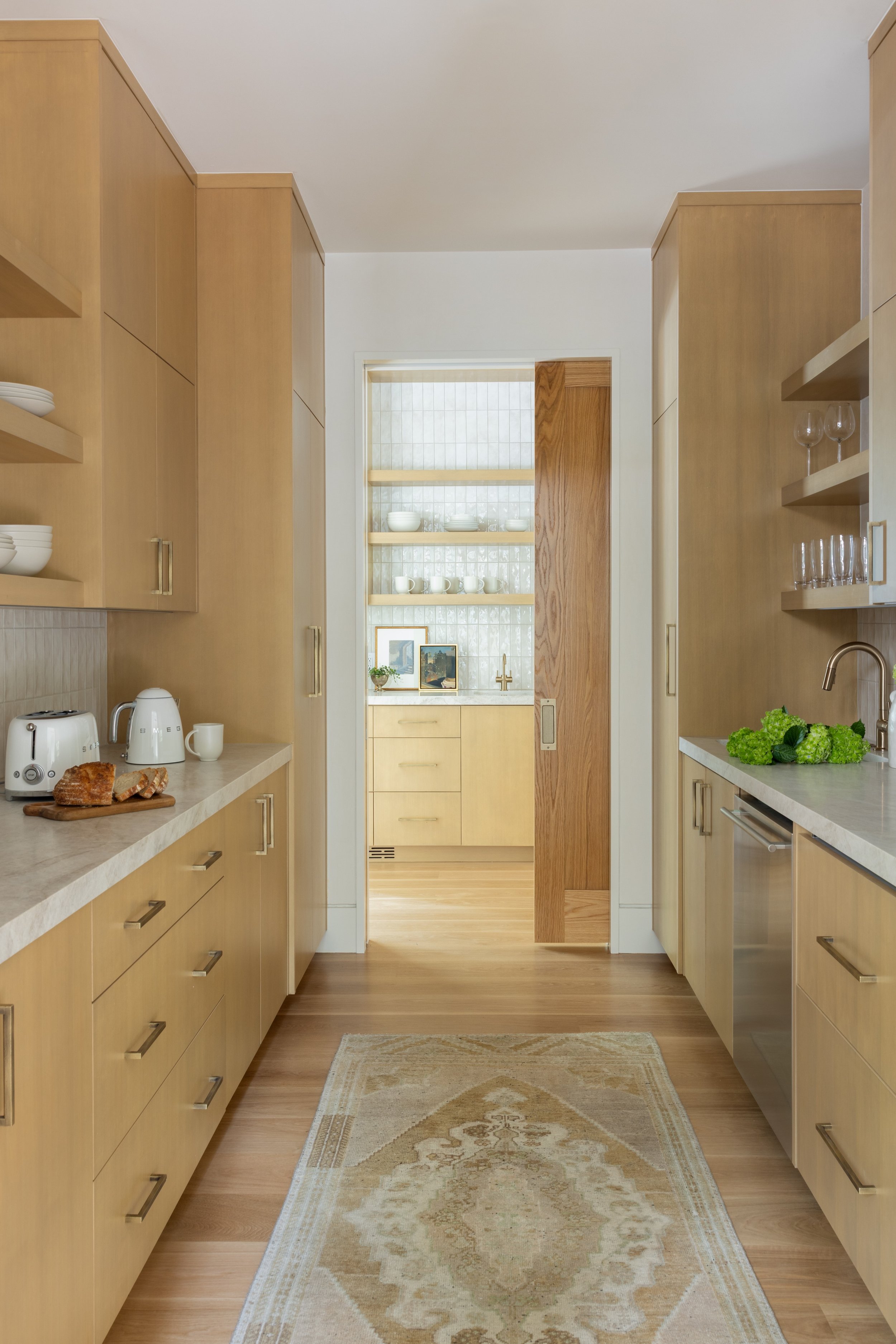 A kitchen with light wooden cabinetry, a white countertop, and a patterned rug on the floor. There are some bread, a toaster, and a kettle on the left counter, and some leafy greens on the right. Open shelving with dishes and glasses is visible.