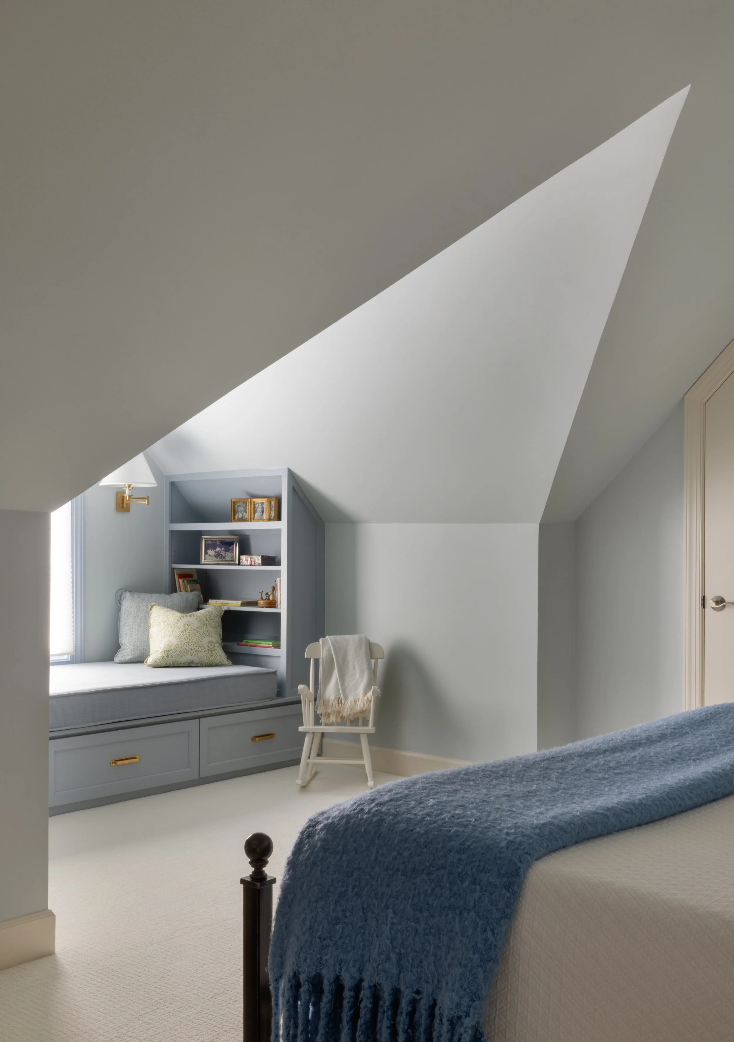 A cozy bedroom with an attic ceiling, a bed with a blue blanket, a window seat with books and pillows, a white chair with a blanket, and a small bookshelf built into the wall.