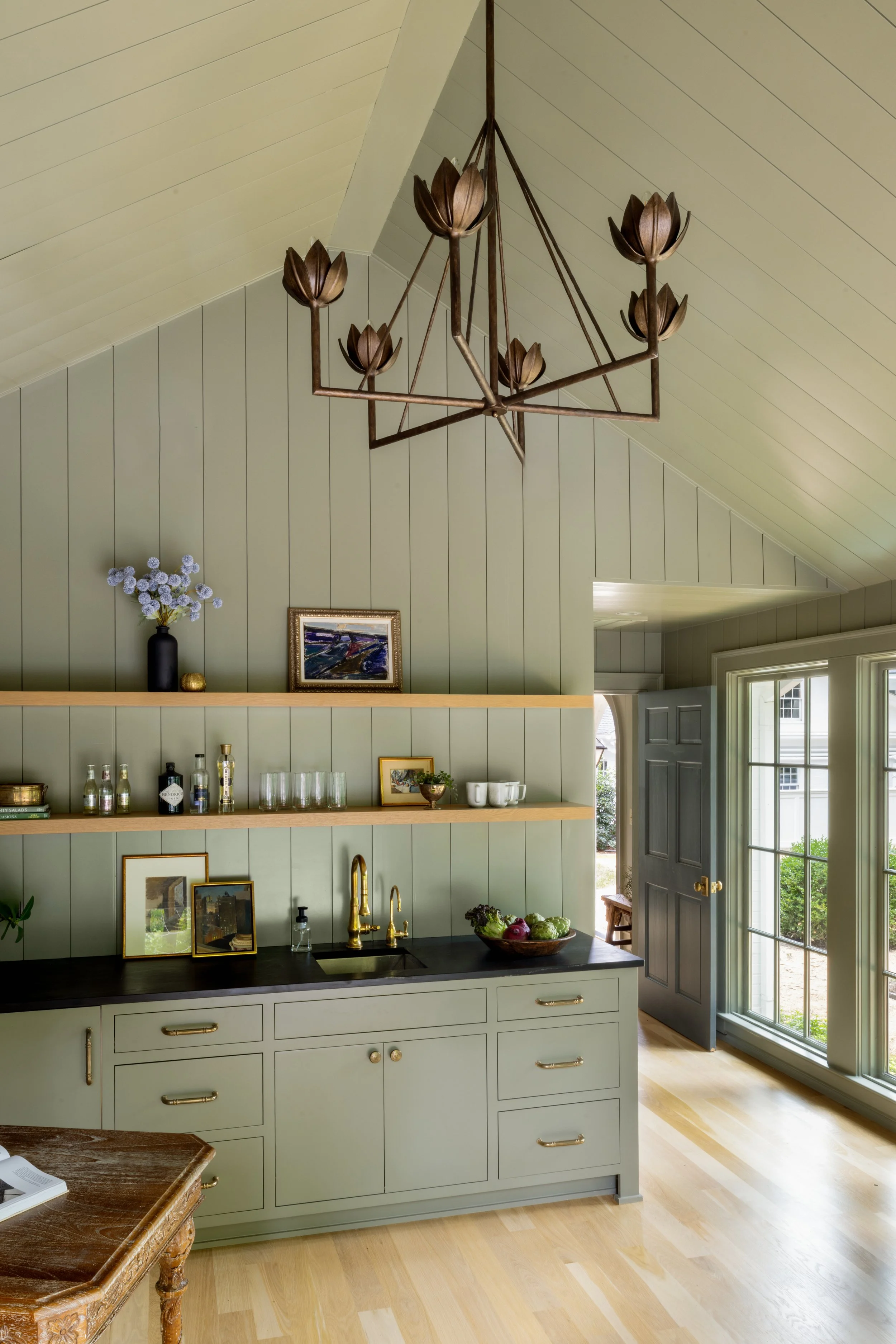 A cozy kitchen with olive green cabinetry, gold hardware, and open wooden shelves displaying bottles, glassware, and pictures. There is a black countertop with a sink and a bowl of vegetables. The tall windows and door let in natural light, and a dec