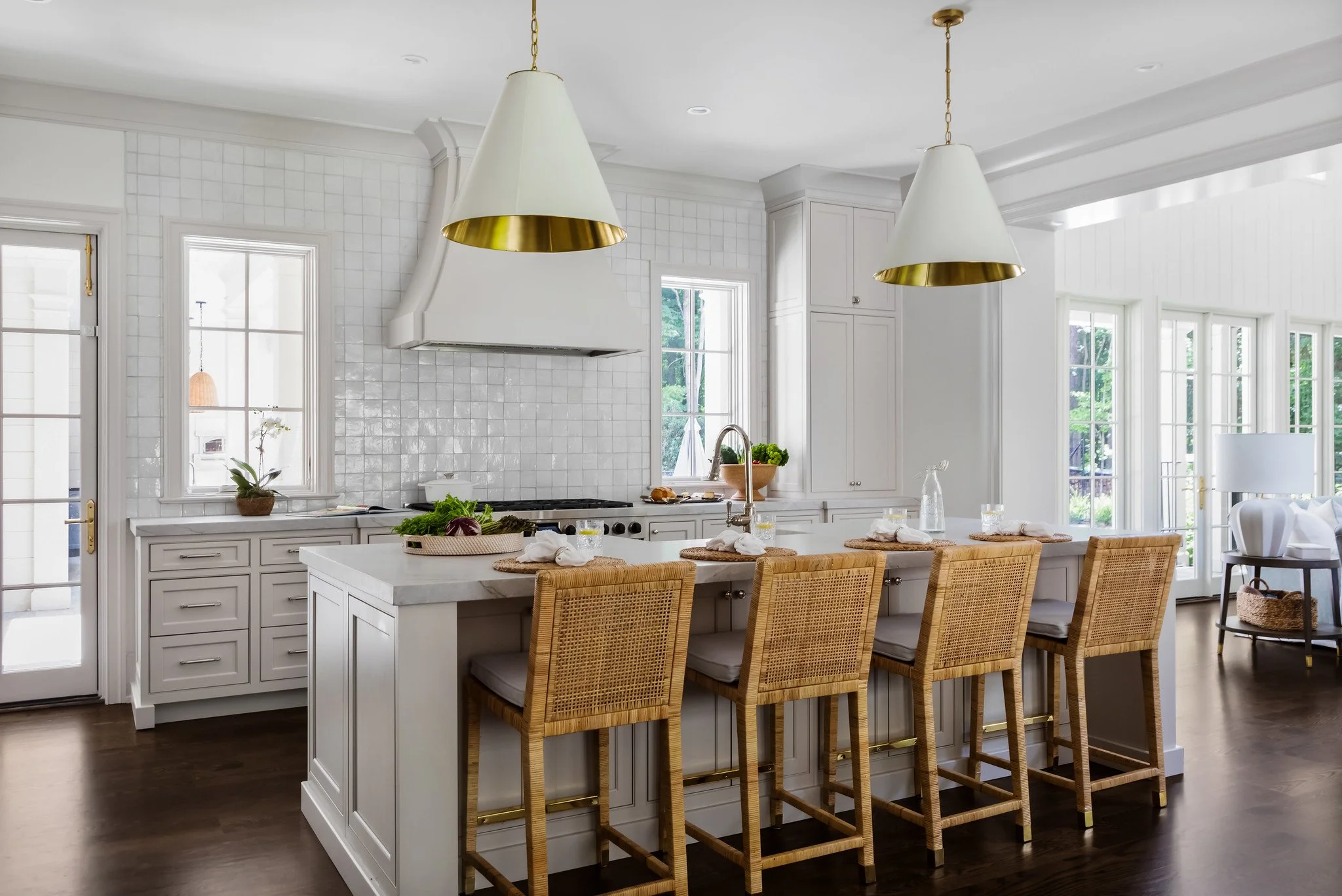 Bright kitchen with white cabinets and walls, dark hardwood flooring, a large island with four wicker chairs, pendant lights, and windows showing greenery outside.