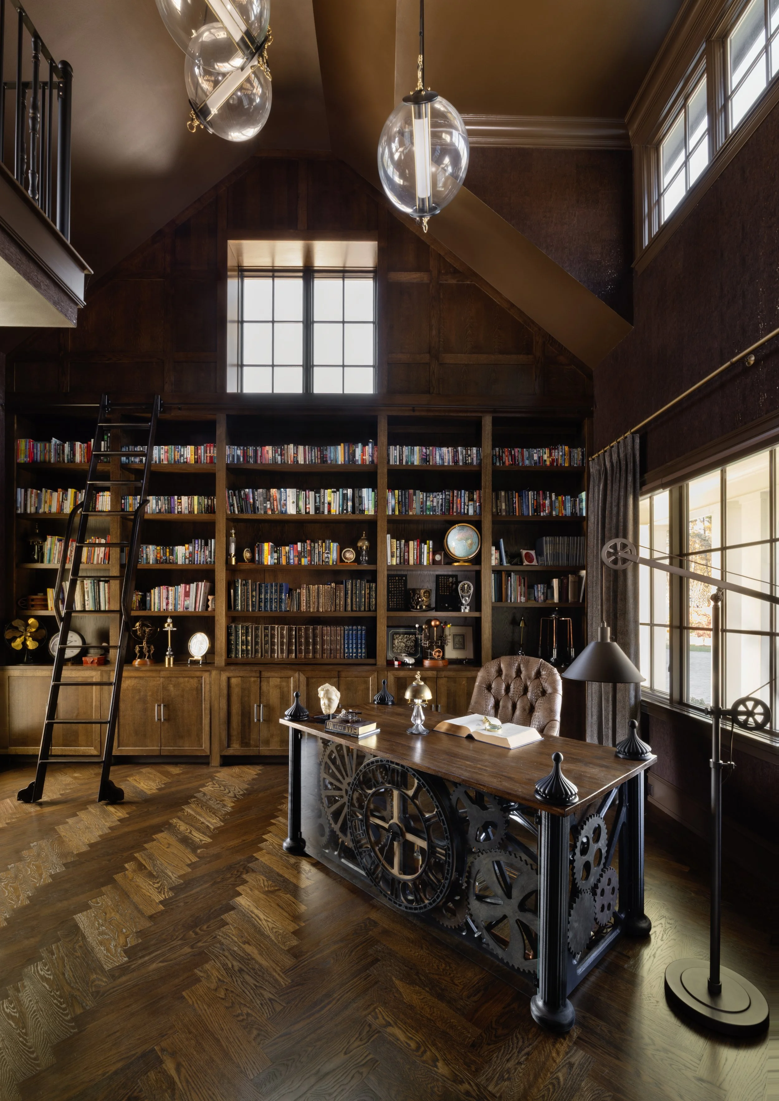 A spacious home library with a large wooden bookshelf filled with books, a wooden desk decorated with vintage items, a tufted leather chair, and large windows letting in natural light. The room has dark wood floors and a high ceiling.