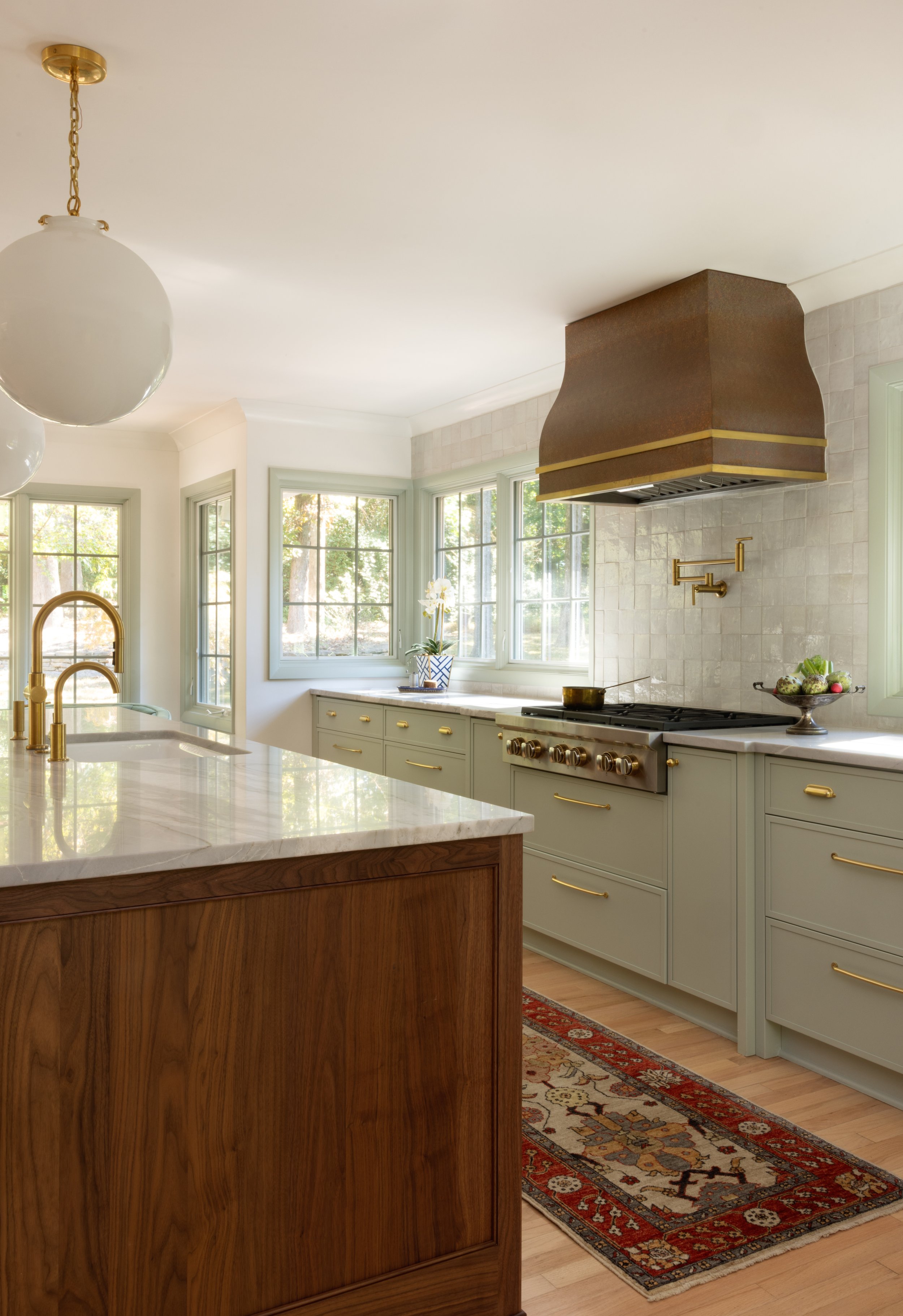 Bright kitchen with light green cabinets, white marble countertops, a gold faucet, a bronze range hood, and windows showing trees outside; decorative rug on wooden floor and a potted plant near the window.
