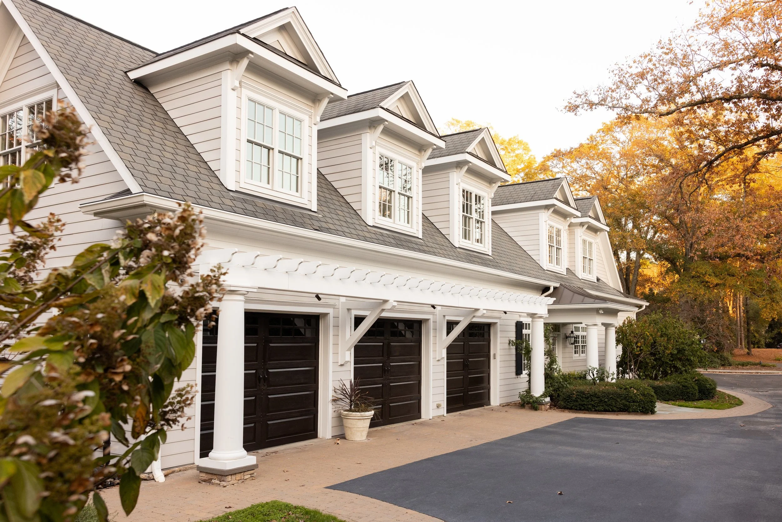 Exterior view of a three-car garage with white siding, black garage doors, and dormer windows, surrounded by trees with fall foliage.