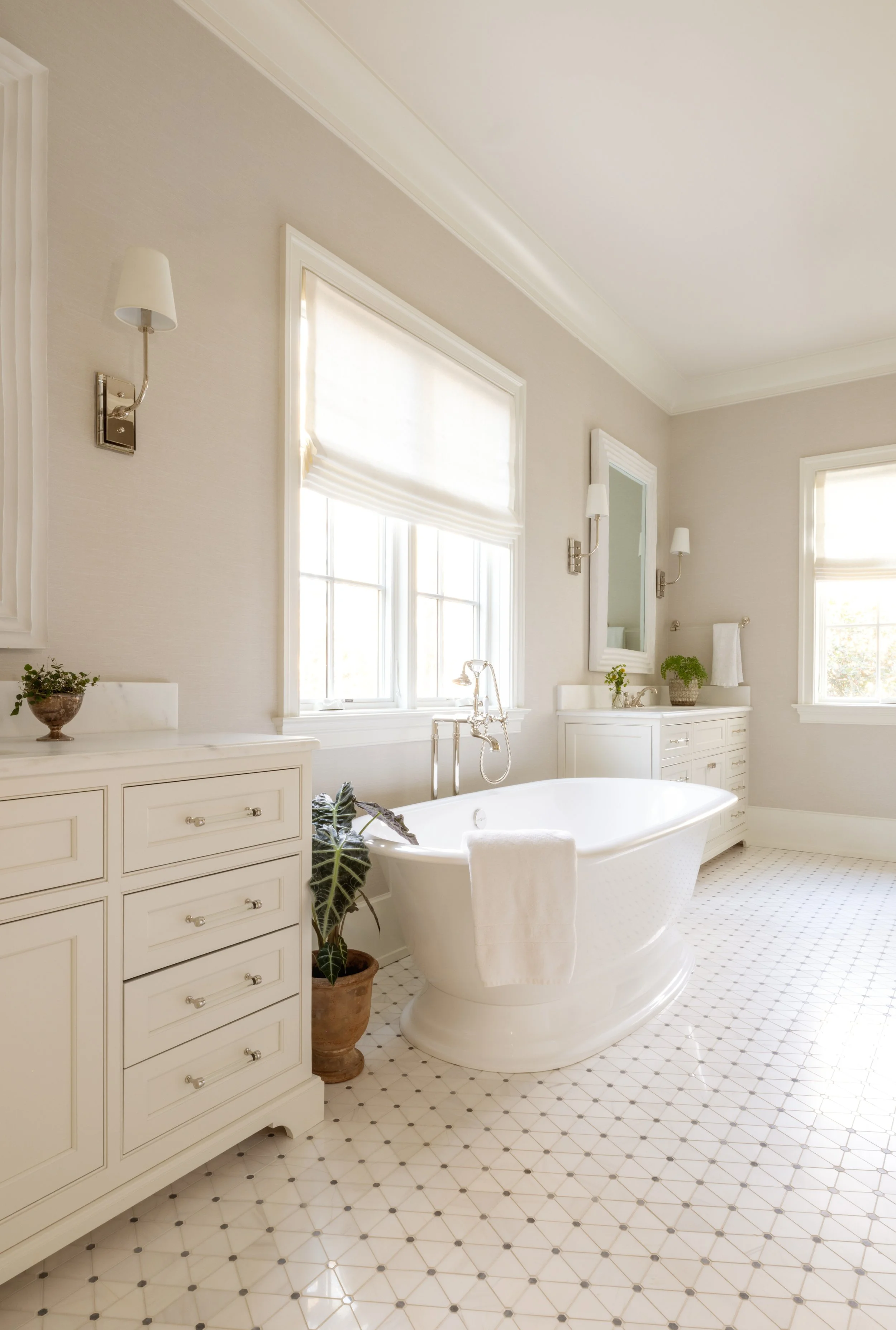 Bright bathroom with a white bathtub, white cabinets, two windows with blinds, and potted plants.