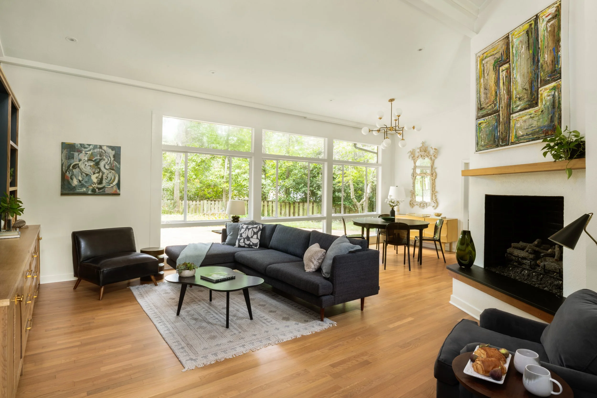 Bright living room with large windows, black sofa, armchair, wooden coffee table, fireplace, dining area, artwork, and hardwood floors.