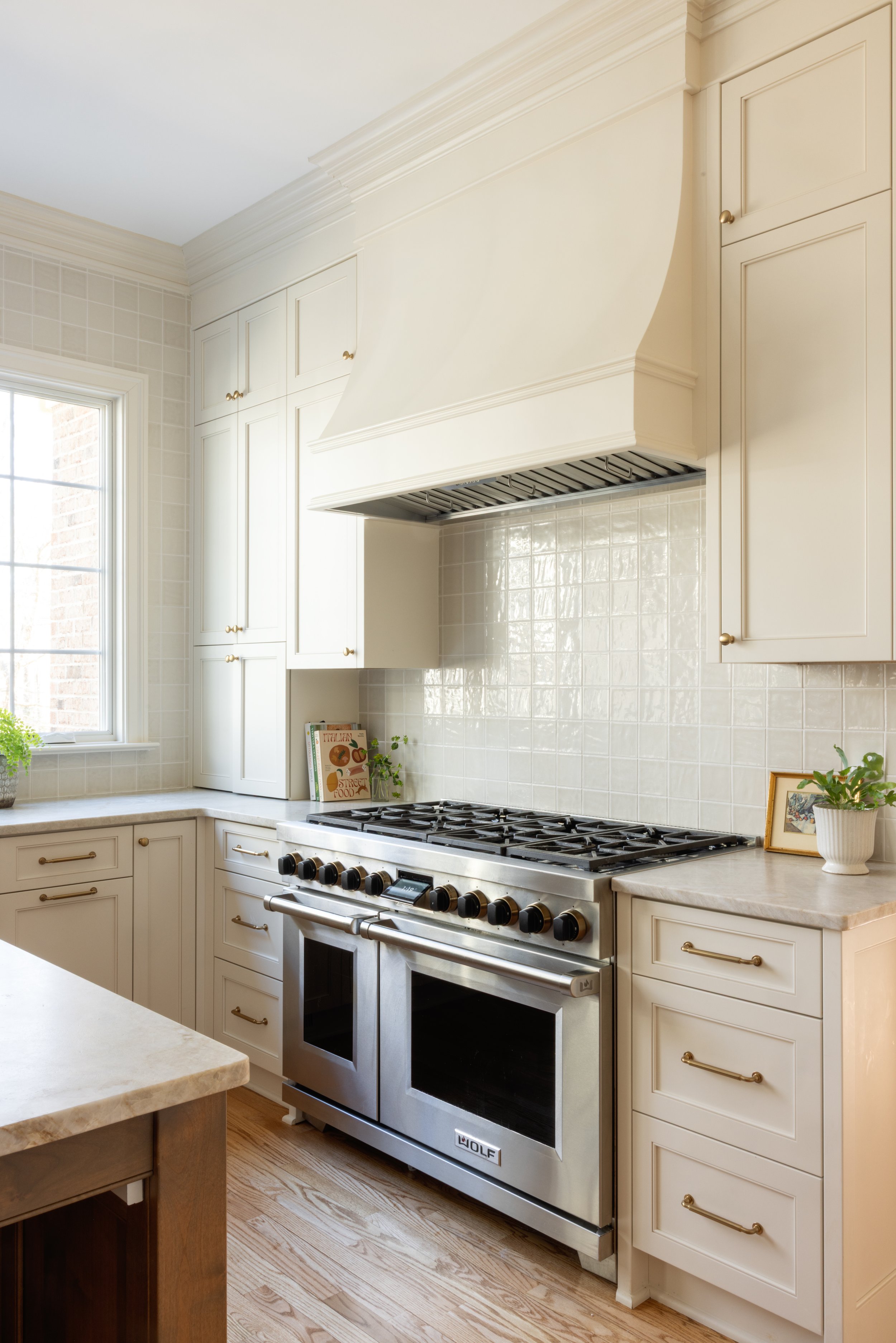 A modern kitchen with white cabinets, a stainless steel oven and stove, beige tiled backsplash, a window with natural light, and small potted plants.