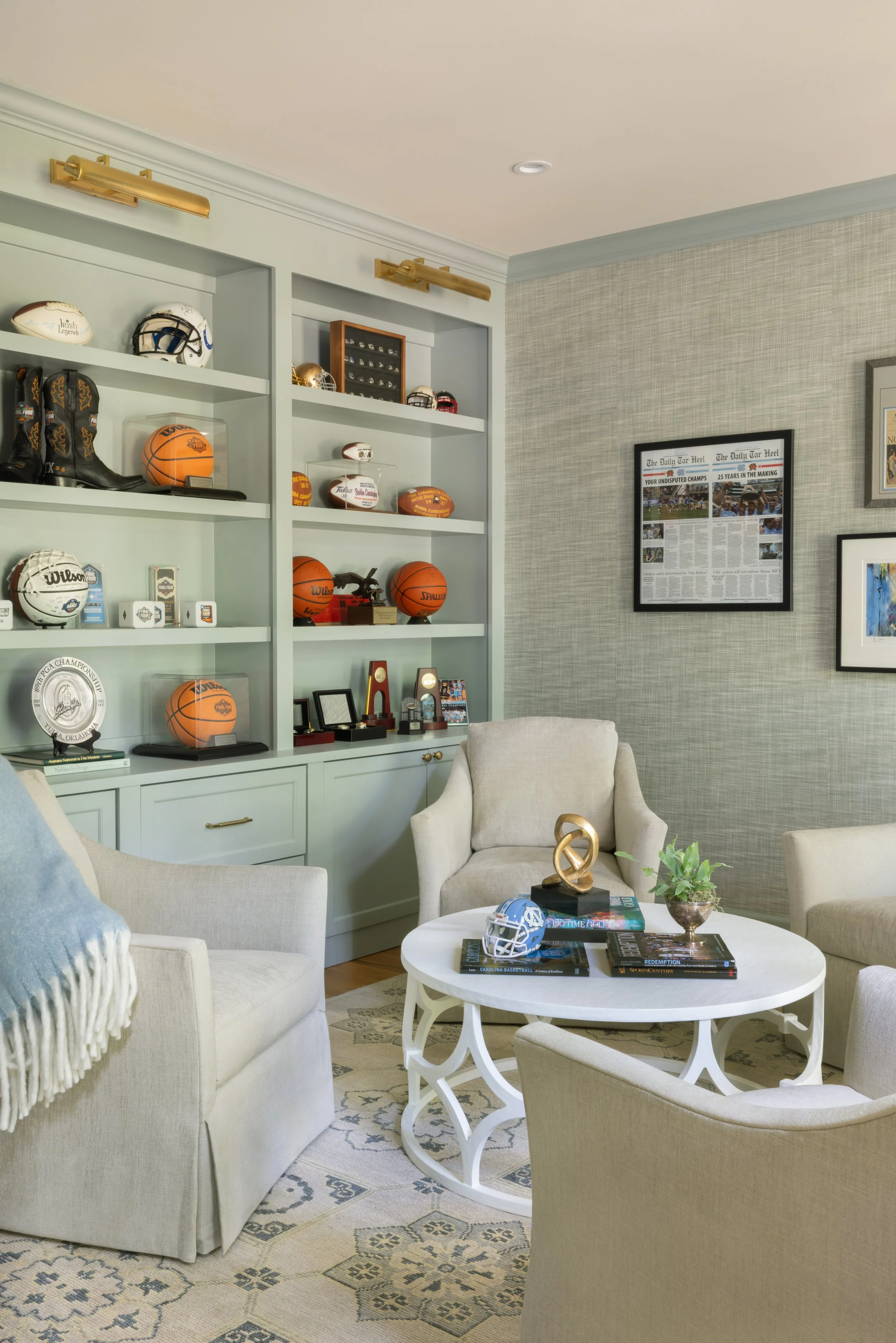 Living room with a built-in shelf displaying sports memorabilia, including footballs, basketballs, a helmet, and awards. There are framed newspaper articles on the wall, and a white coffee table with books, a small plant, and decorative items. Severa
