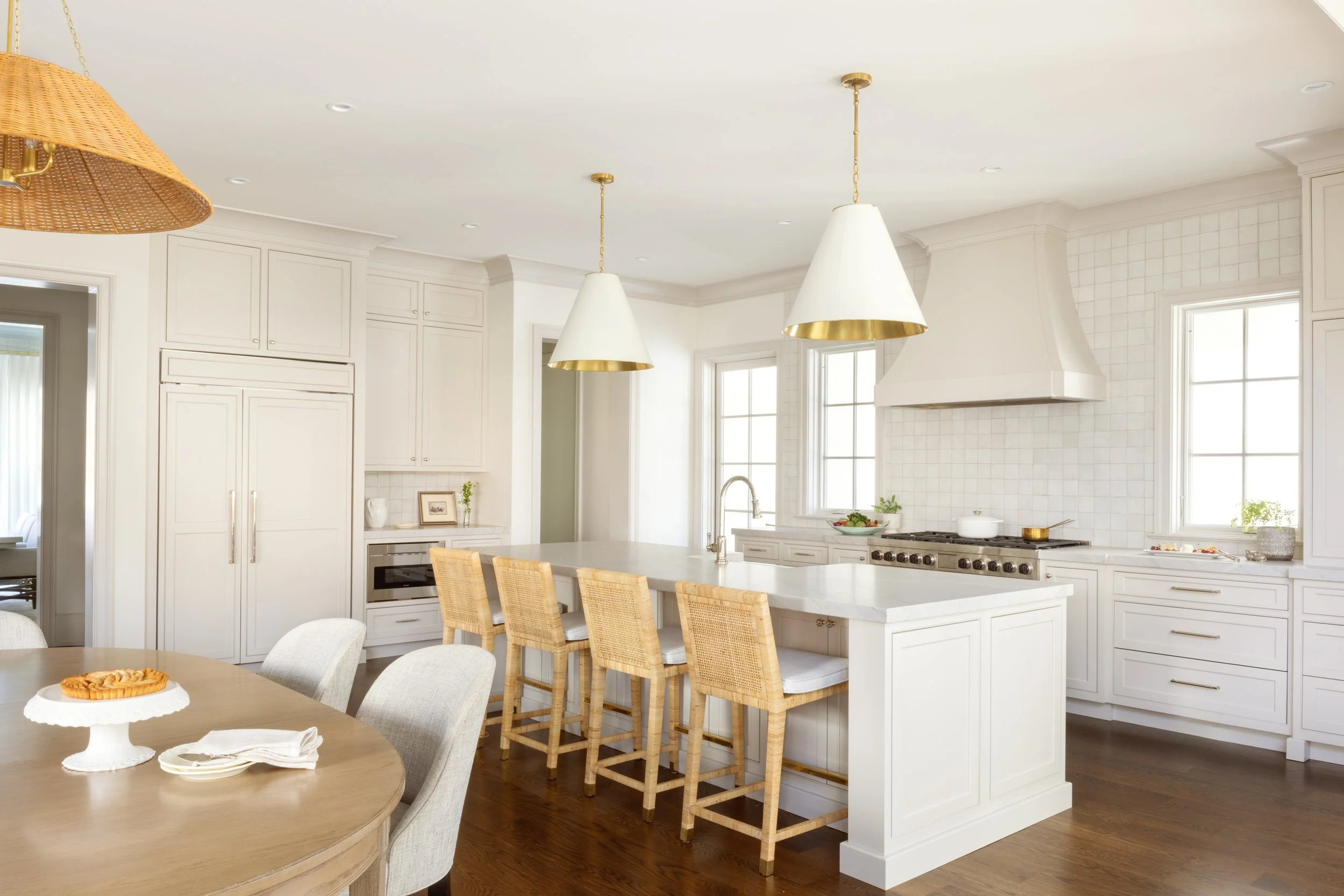 Bright white kitchen with large island, four barstools, pendant lights, and a wood dining table with chairs, decorated with a cookie and napkin, hardwood floors.