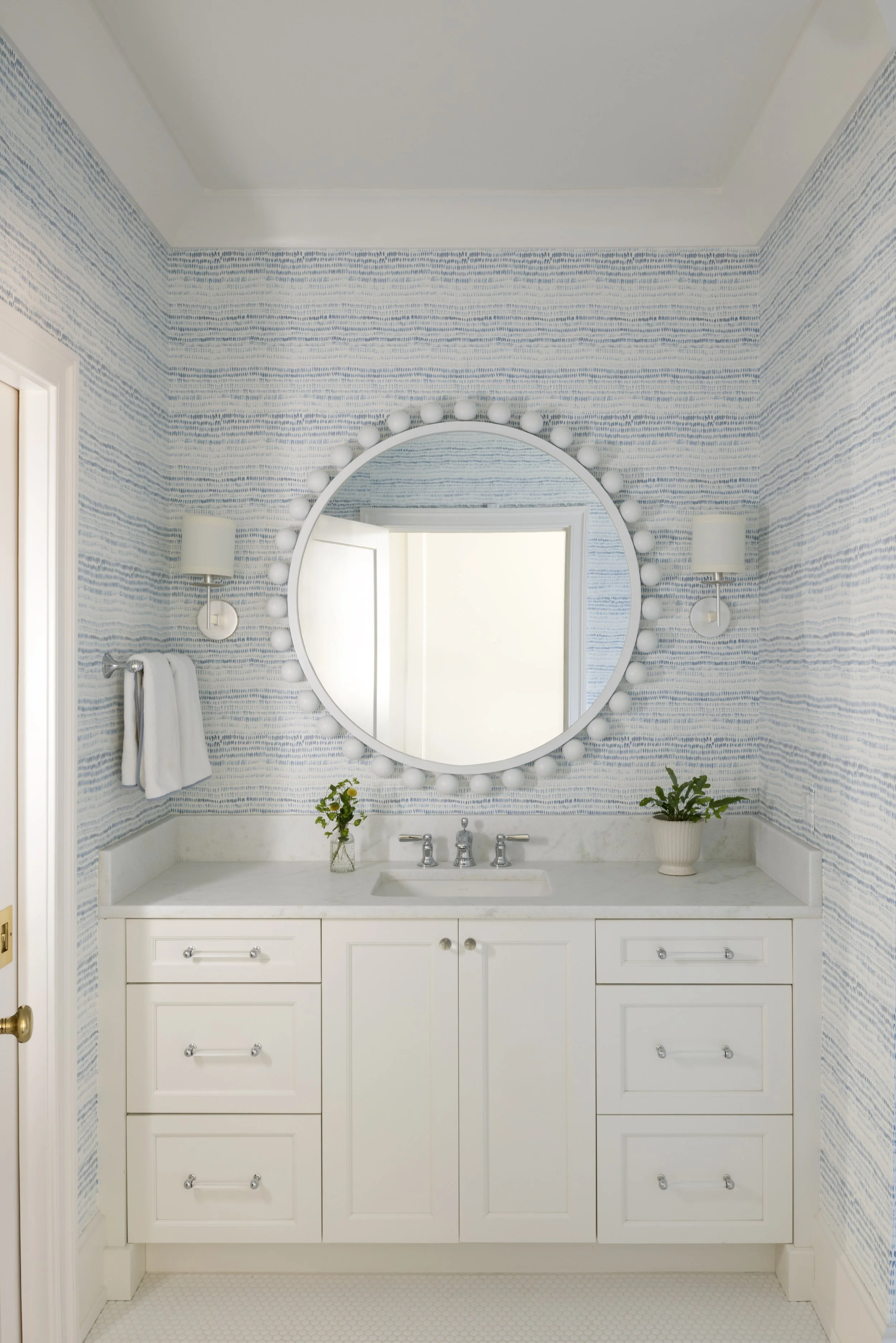 White bathroom vanity with a marble countertop, two potted plants, and a large round mirror with globe bulbs around its perimeter. Blue patterned wallpaper and wall-mounted light fixtures on each side of the mirror.